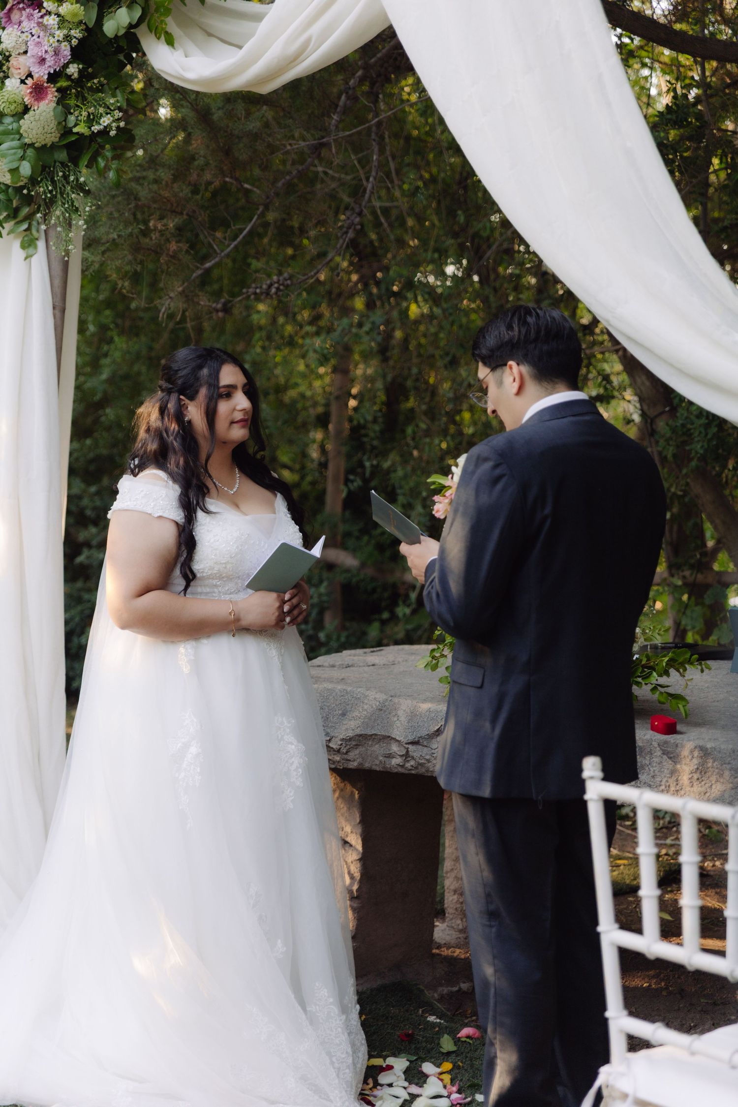 Bride and groom reading wedding vows under white draped floral arch in garden ceremony.