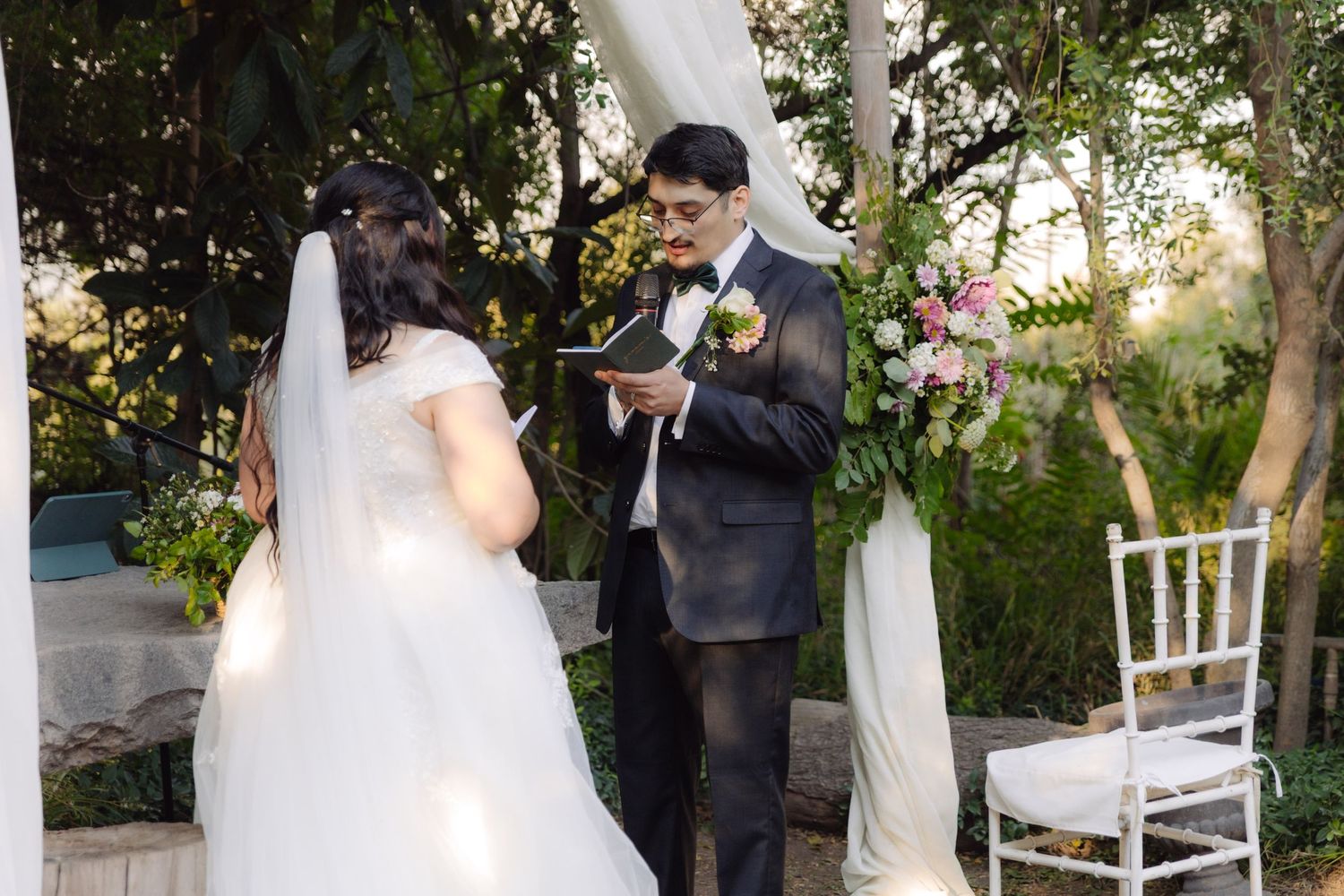 Groom reading wedding vows to bride under white draped arch with floral arrangements.