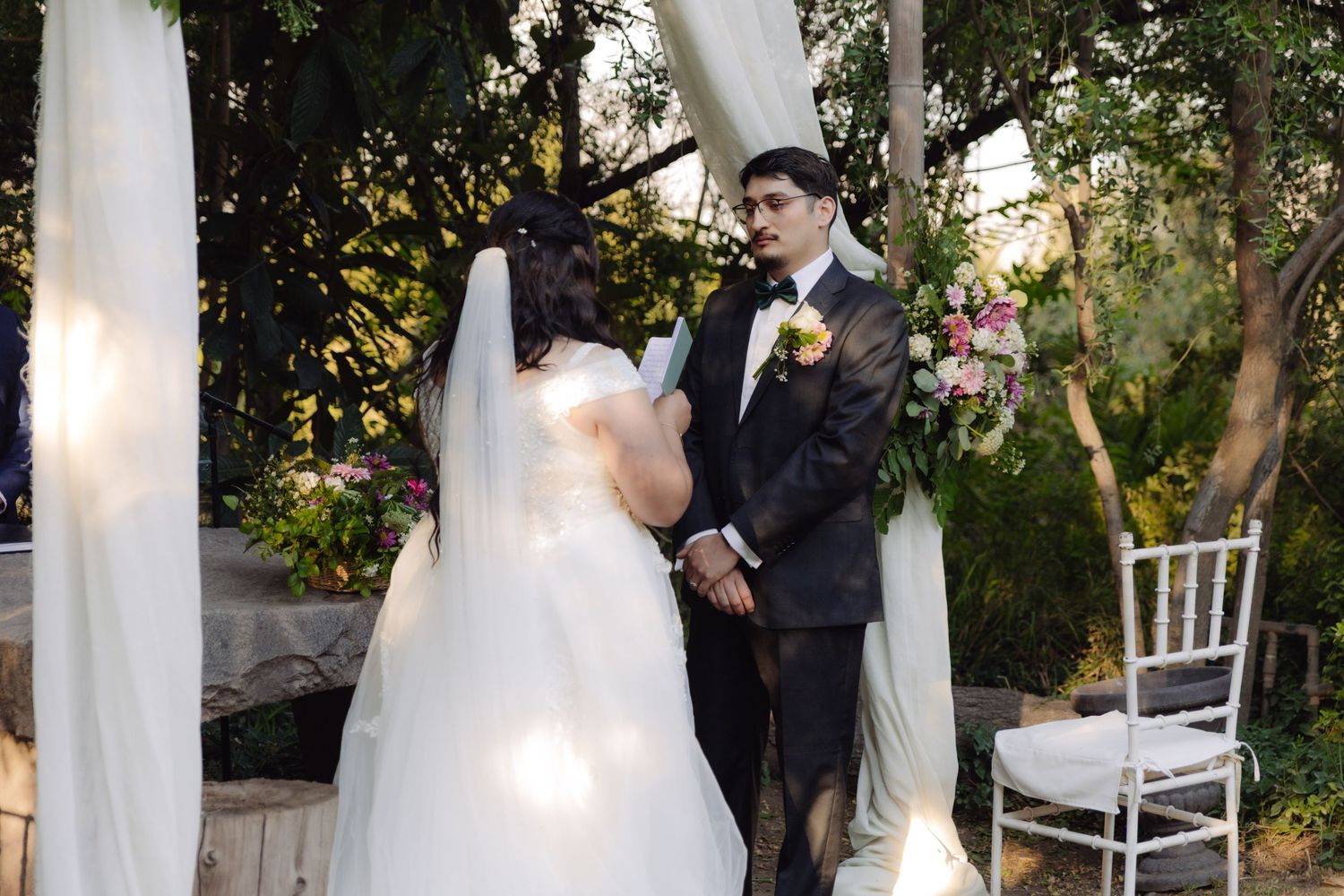 Bride and groom facing each other under white draped wedding arch in garden setting.
