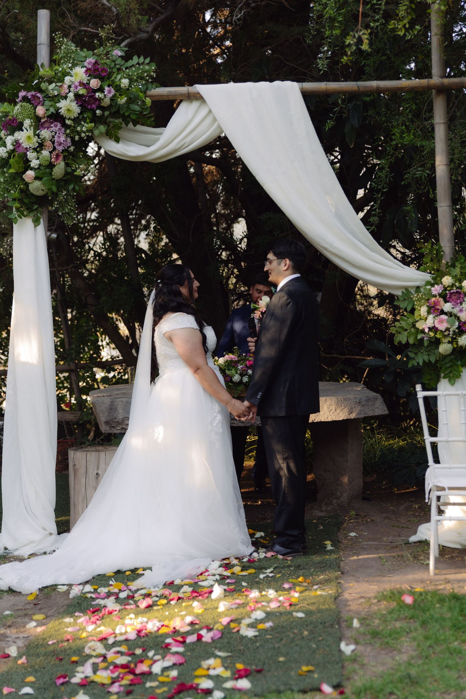 Bride and groom hold hands at outdoor wedding altar adorned with white drapes and floral arrangements on petal-lined aisle.