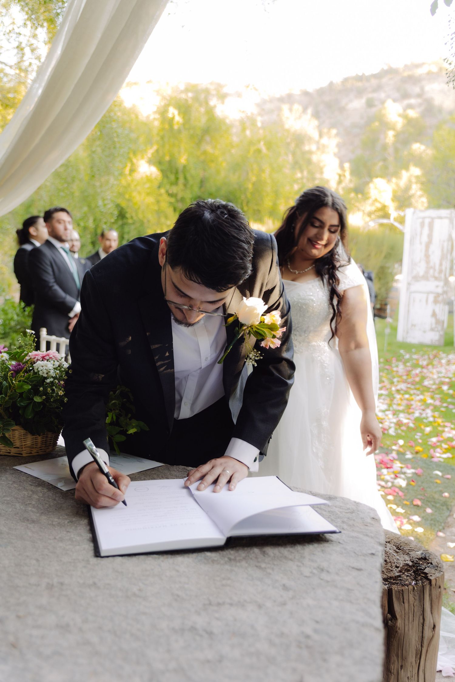 Groom in black suit signs wedding registry book as smiling bride in white dress watches during outdoor ceremony.