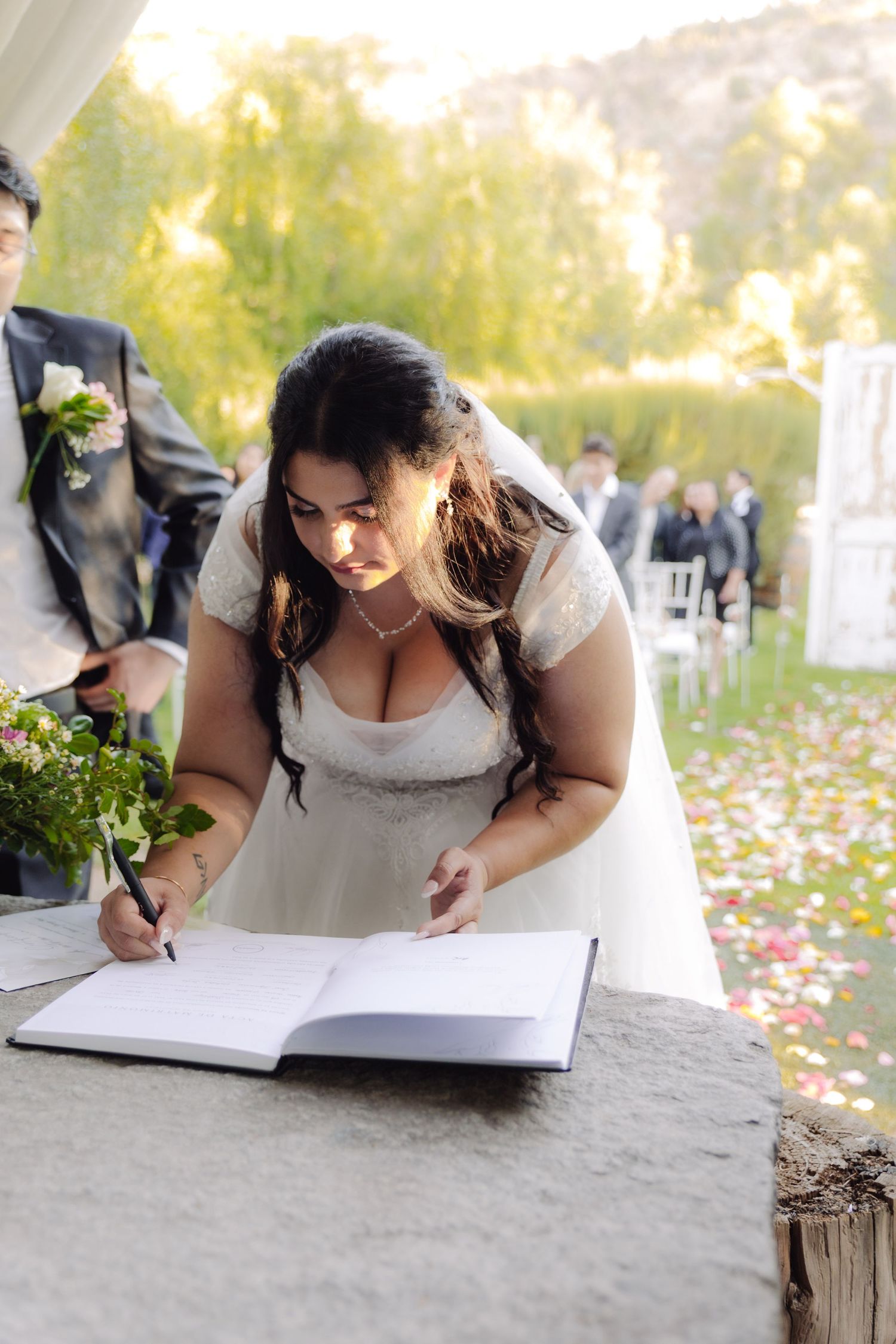 Bride in white lace wedding dress signs marriage registry book at outdoor ceremony with colorful petals on ground.
