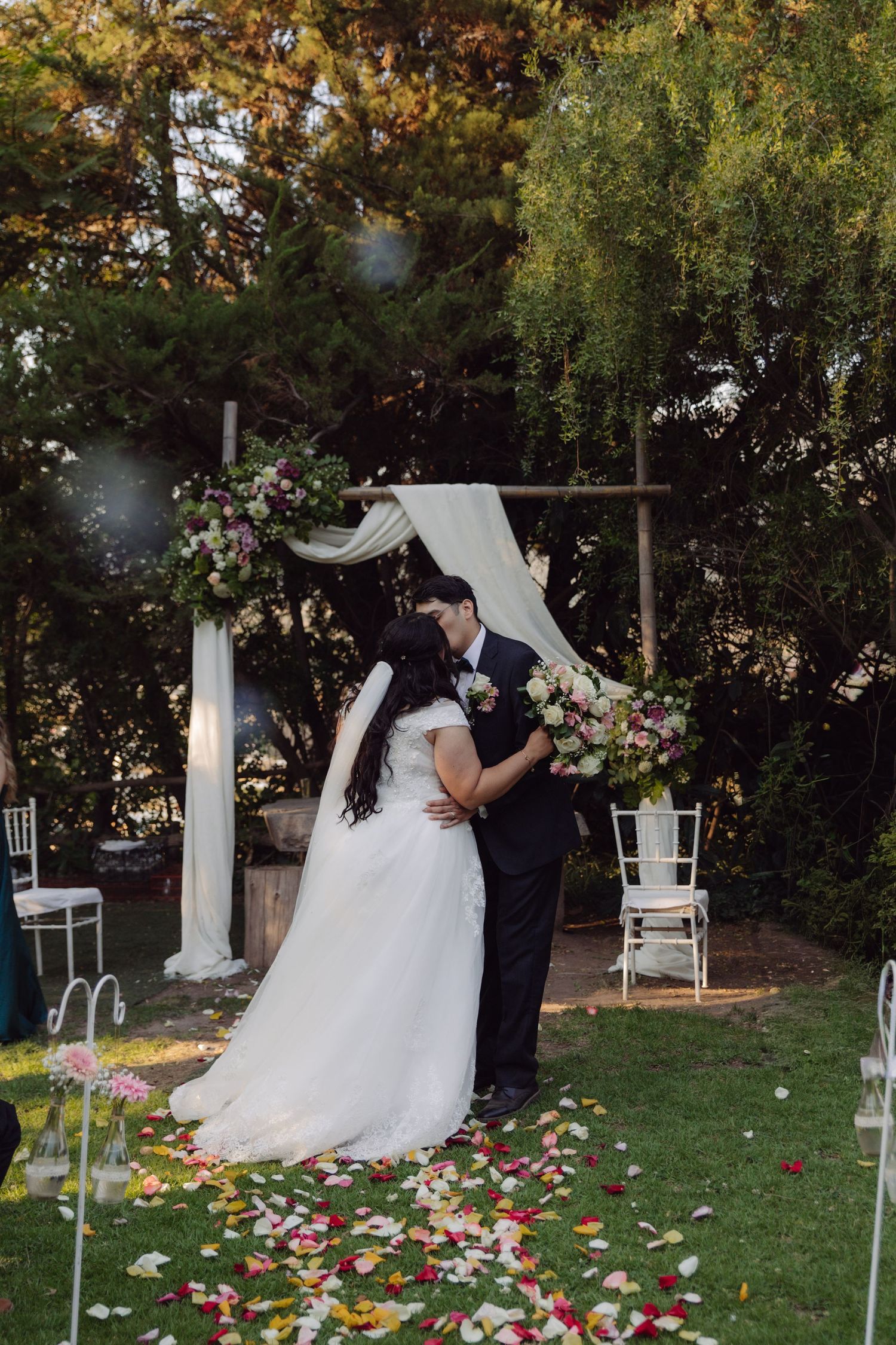 Newlyweds share first kiss under rustic wooden arch with white drapes and flowers on petal-covered garden aisle.