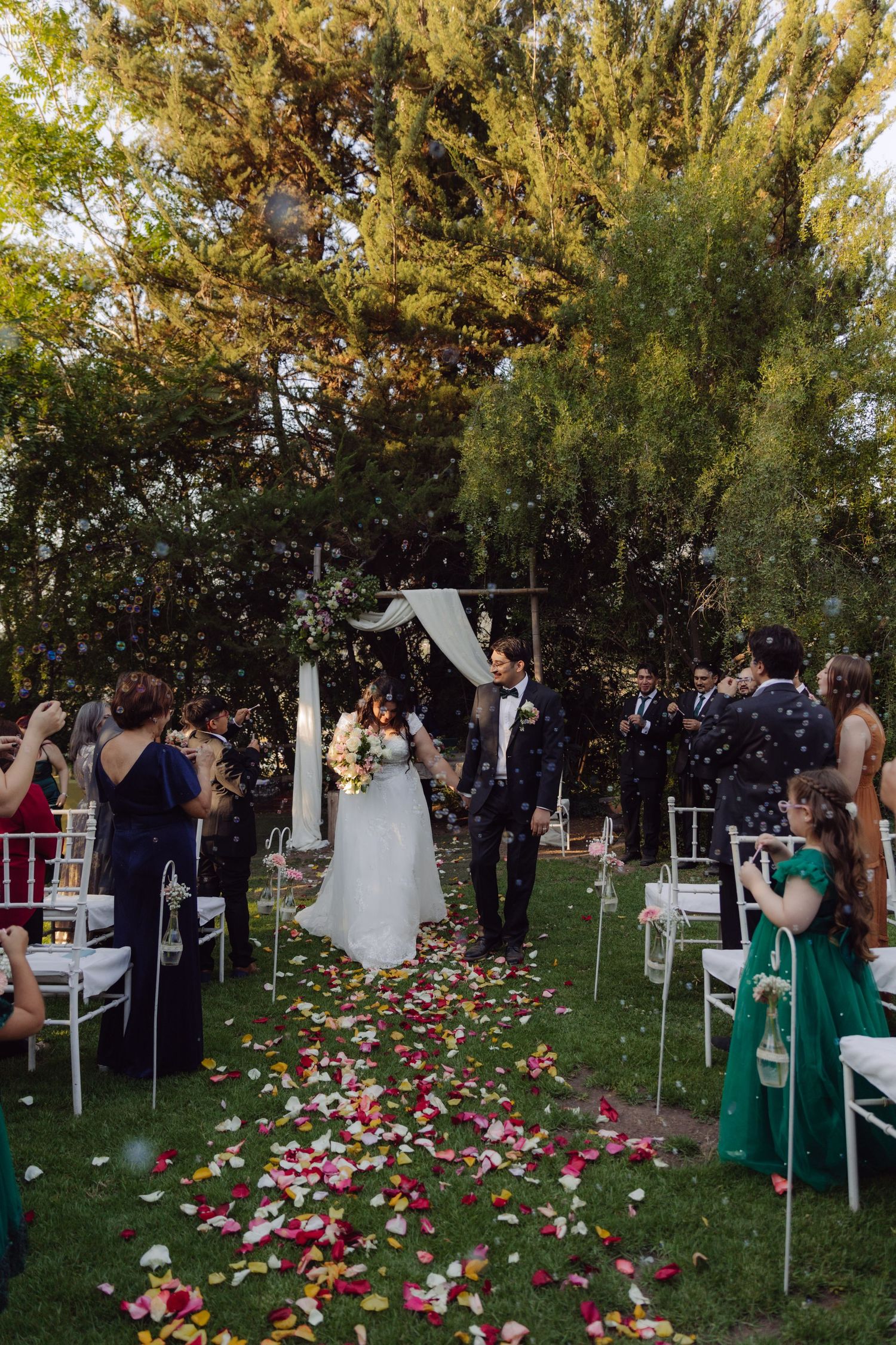 Newlyweds walk joyfully down petal-lined aisle as guests celebrate with confetti at outdoor garden wedding ceremony.