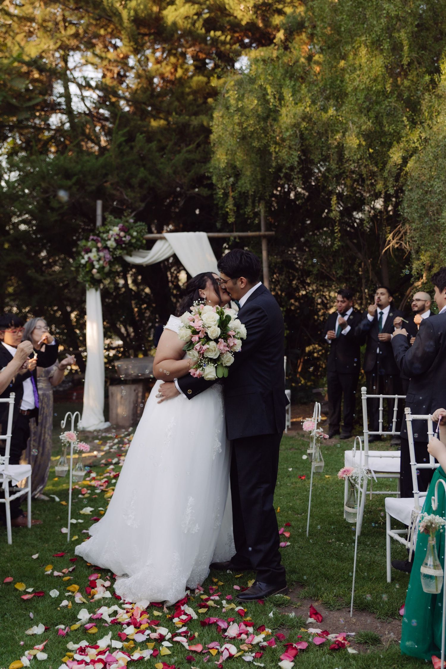 Newlyweds share romantic kiss on petal-strewn garden aisle as guests applaud during outdoor wedding ceremony.