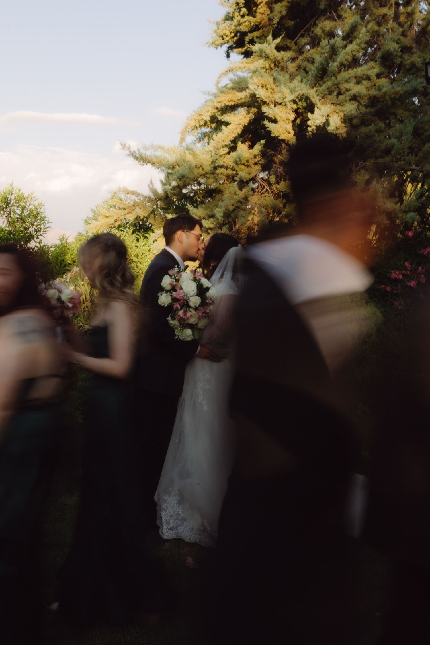 Bride and groom share intimate moment among lush garden greenery while guests move around them after ceremony.
