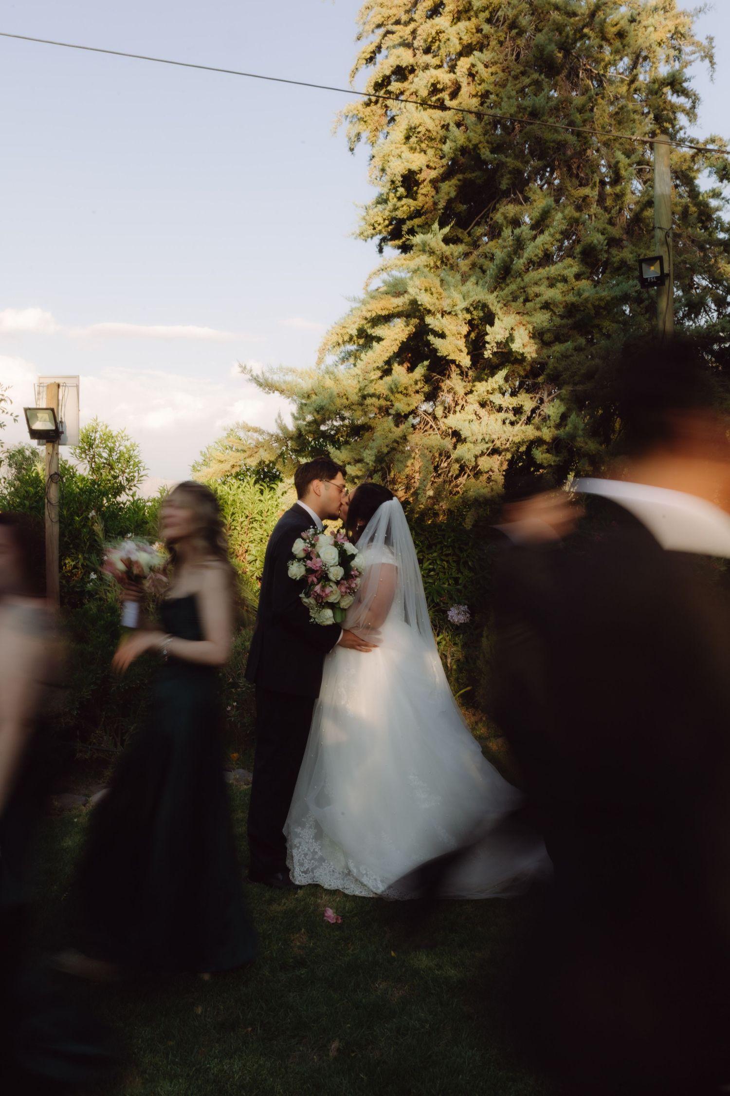 Bride and groom share a kiss in lush garden setting while wedding guests move around them.