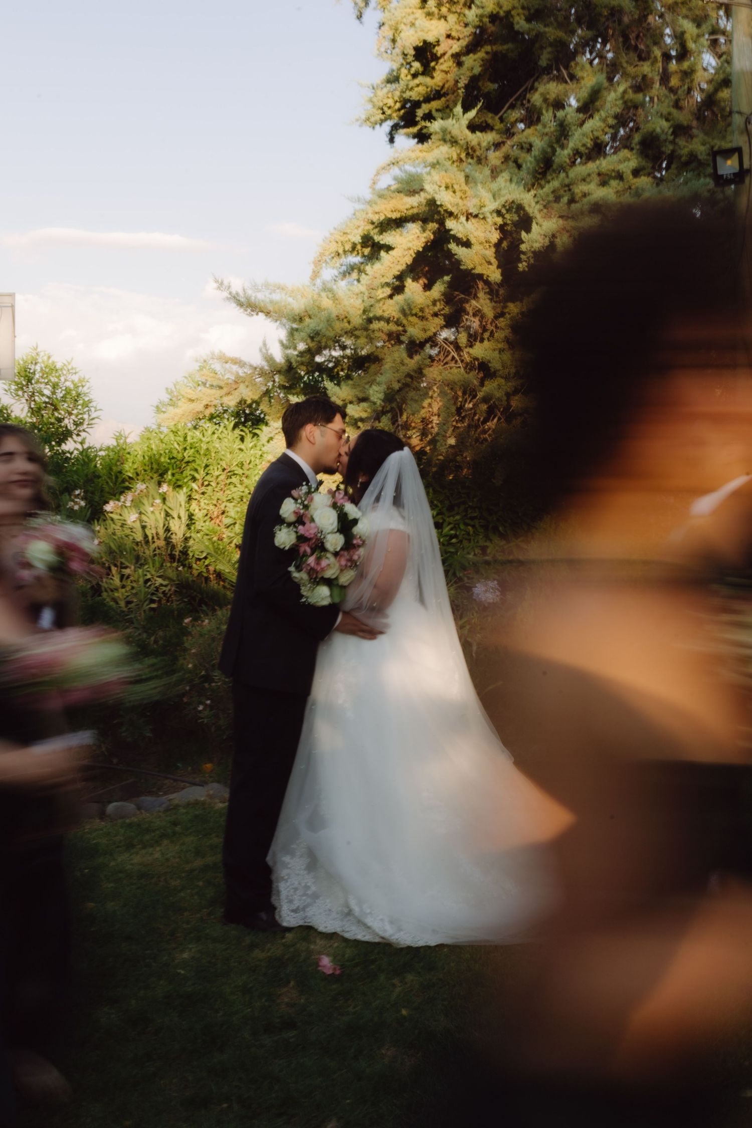 Bride holding pink and white bouquet kisses groom in black suit in beautiful garden setting.