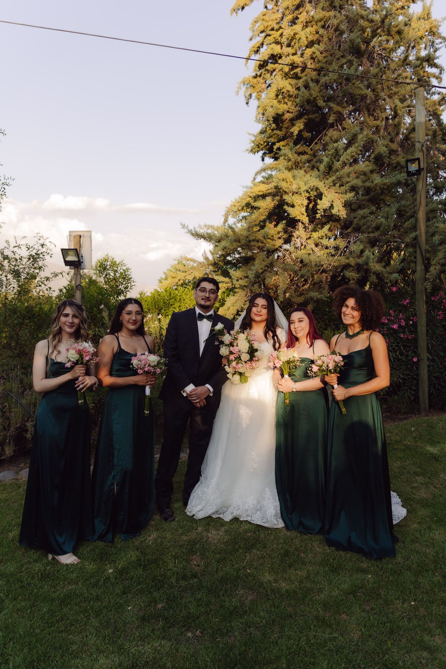 Bride and groom pose with four bridesmaids in elegant dark green satin gowns in garden setting.