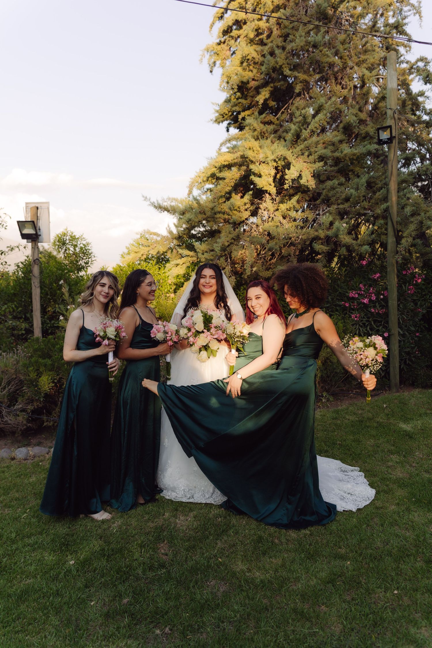 Bride laughs with four bridesmaids in dark green satin dresses holding pink bouquets in garden.