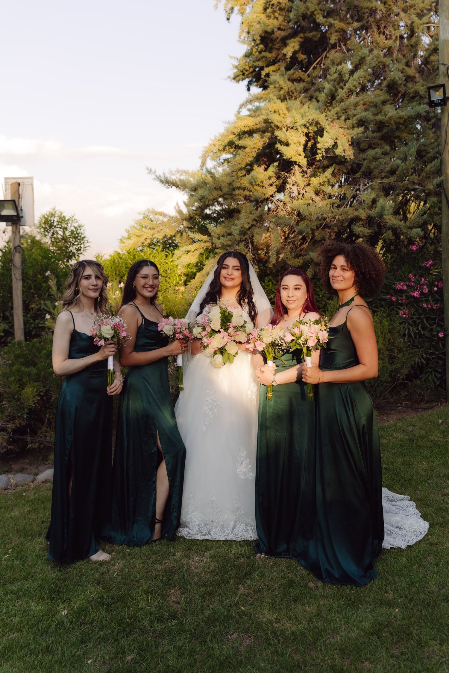 Bride in white gown with four bridesmaids in emerald green dresses holding pink bouquets outdoors.
