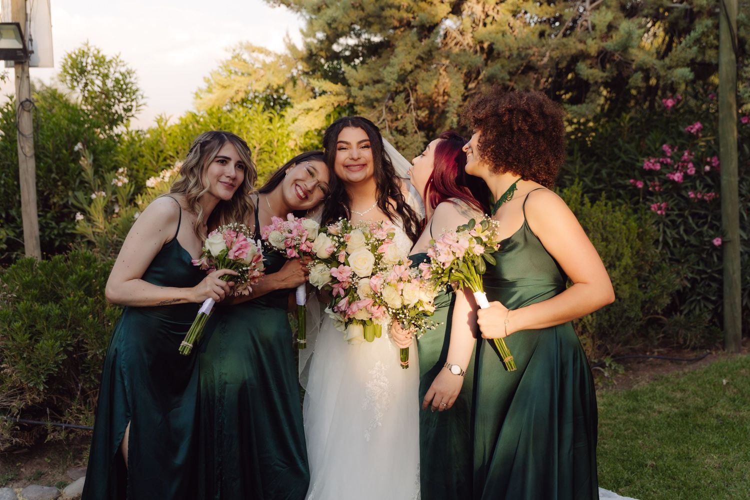 Bride and bridesmaids in green dresses laughing together while holding floral bouquets at wedding.