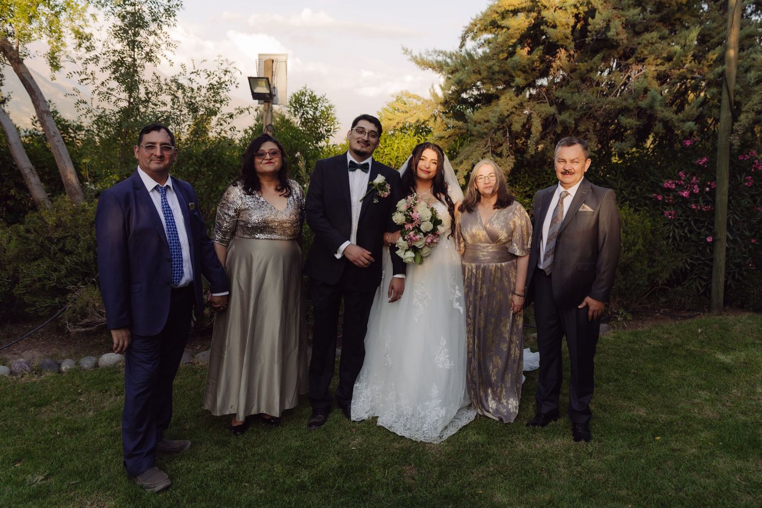 Wedding couple poses with family members in formal attire in a garden at golden hour.