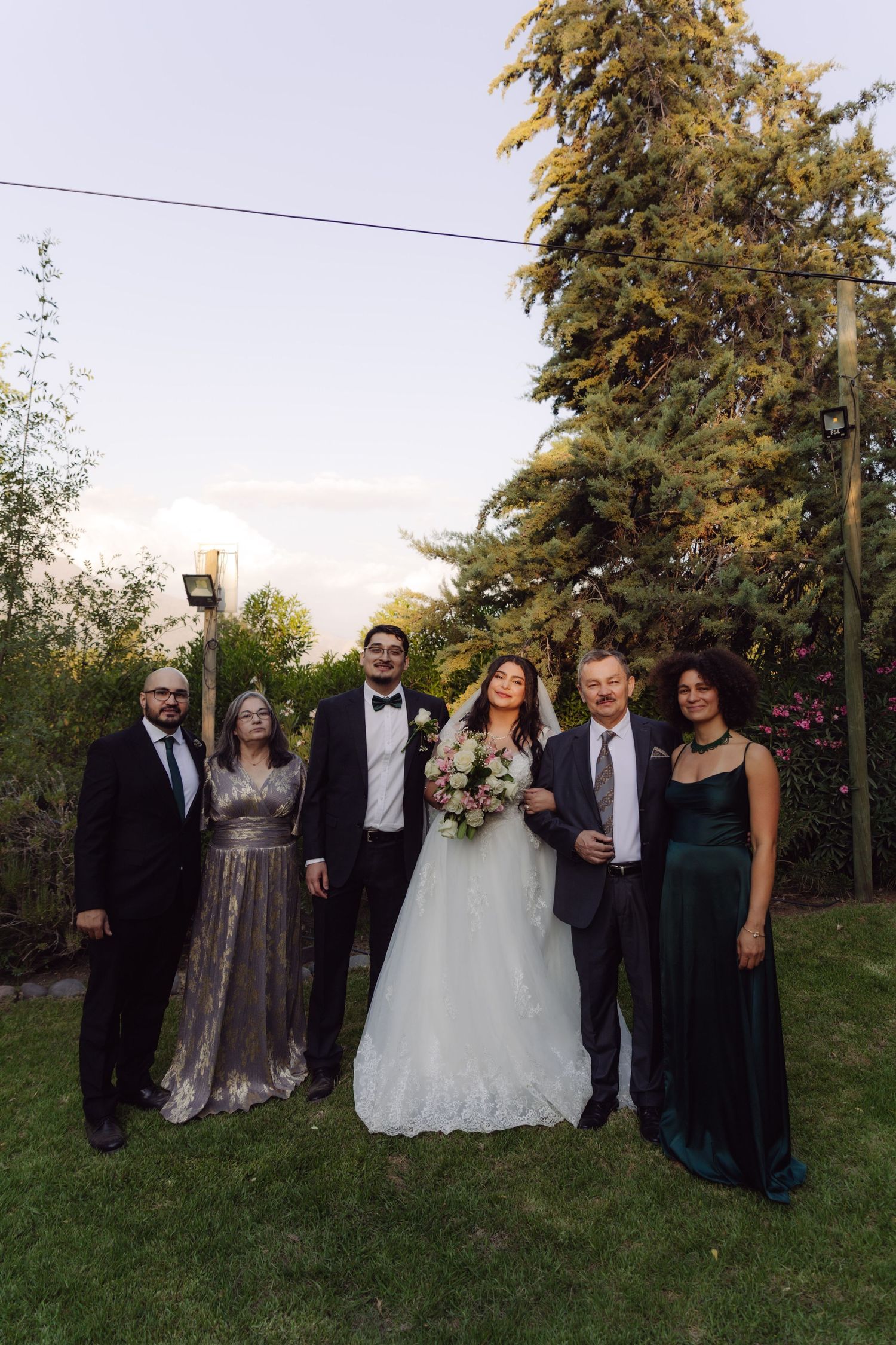 Bride and groom stand with family group in formal wear beside tall evergreen tree outdoors.