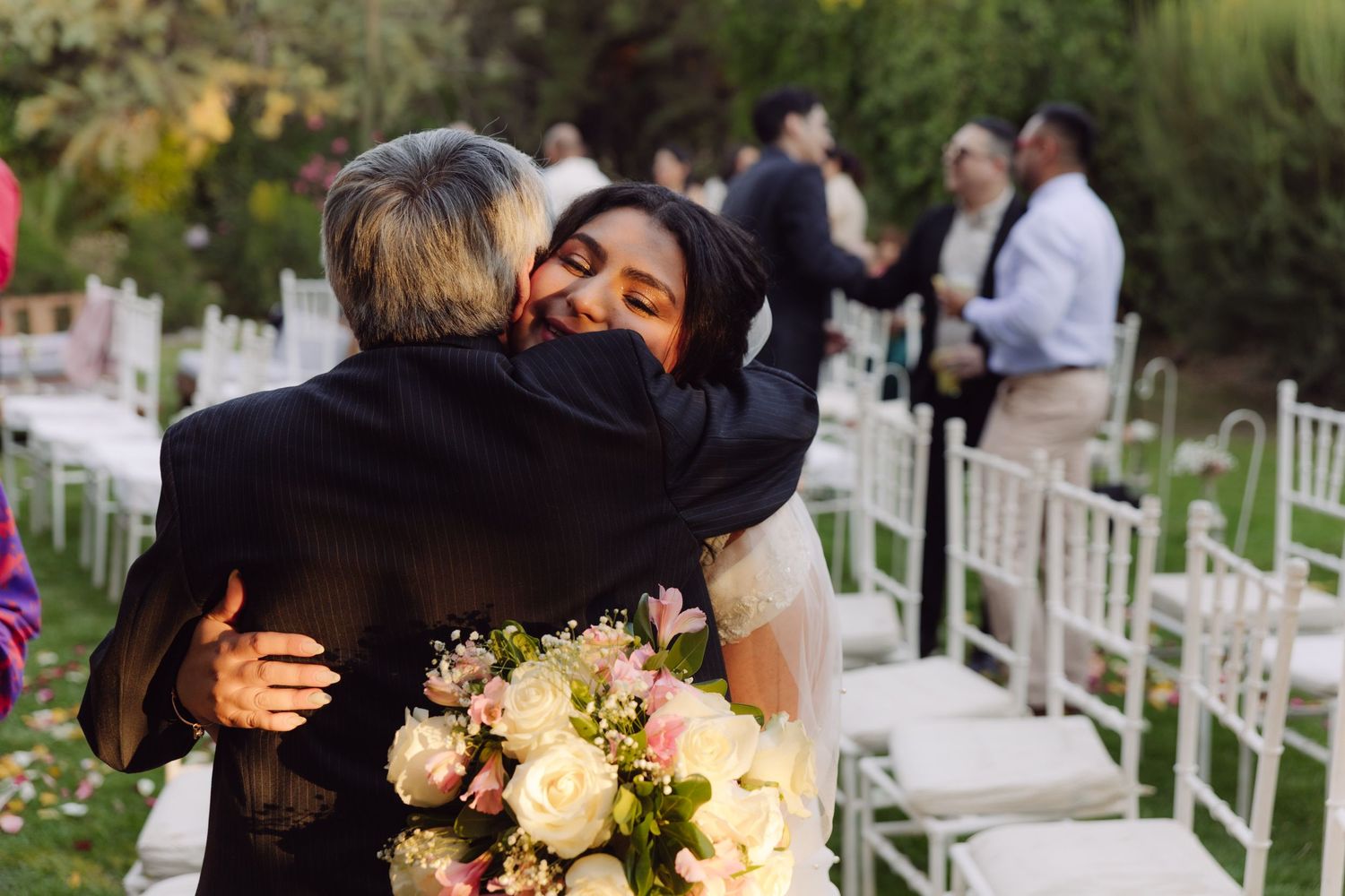 Bride in white dress emotionally hugs guest while holding pink bouquet at outdoor wedding ceremony.