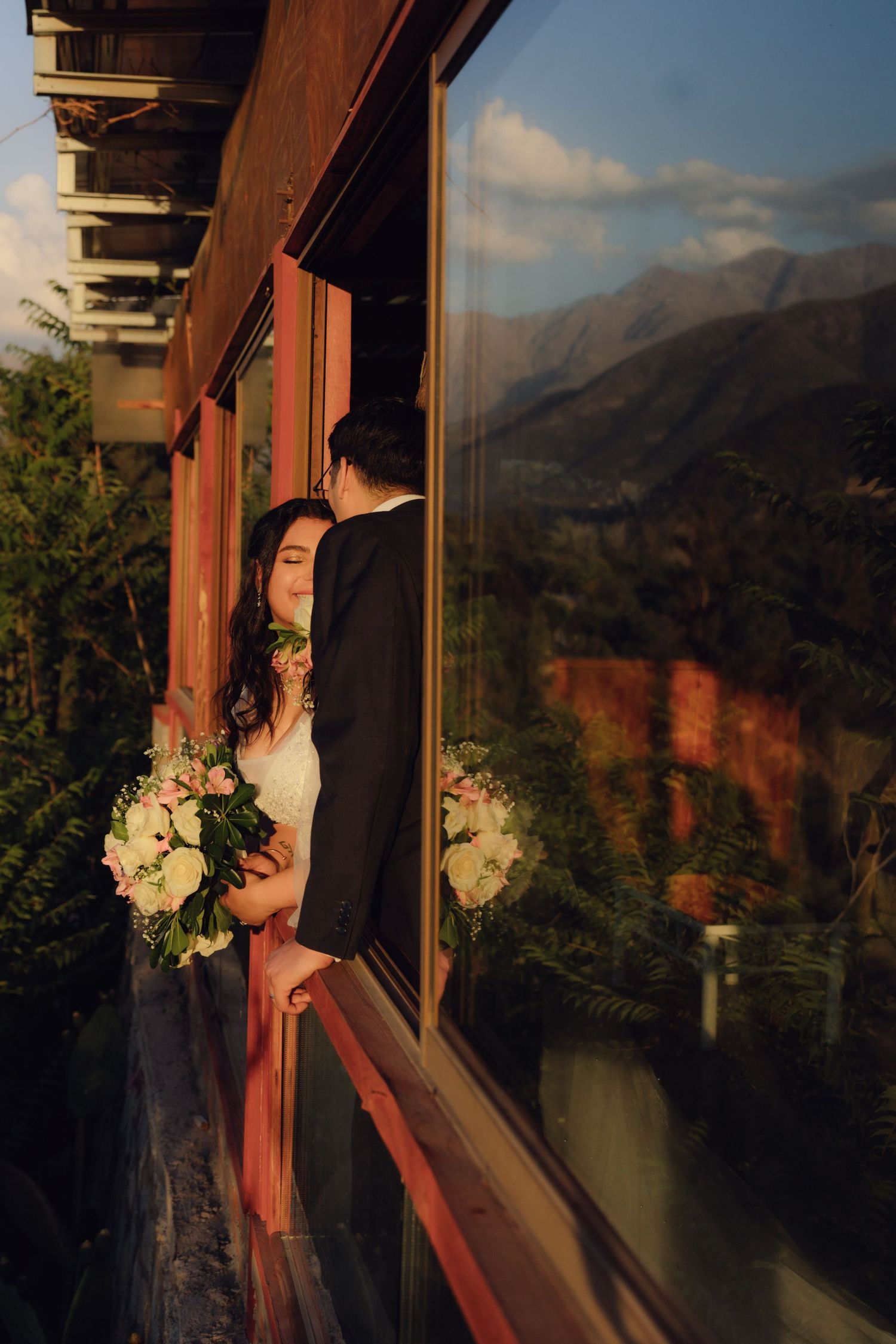 Bride and groom share intimate moment on wooden balcony with stunning mountain landscape reflection.