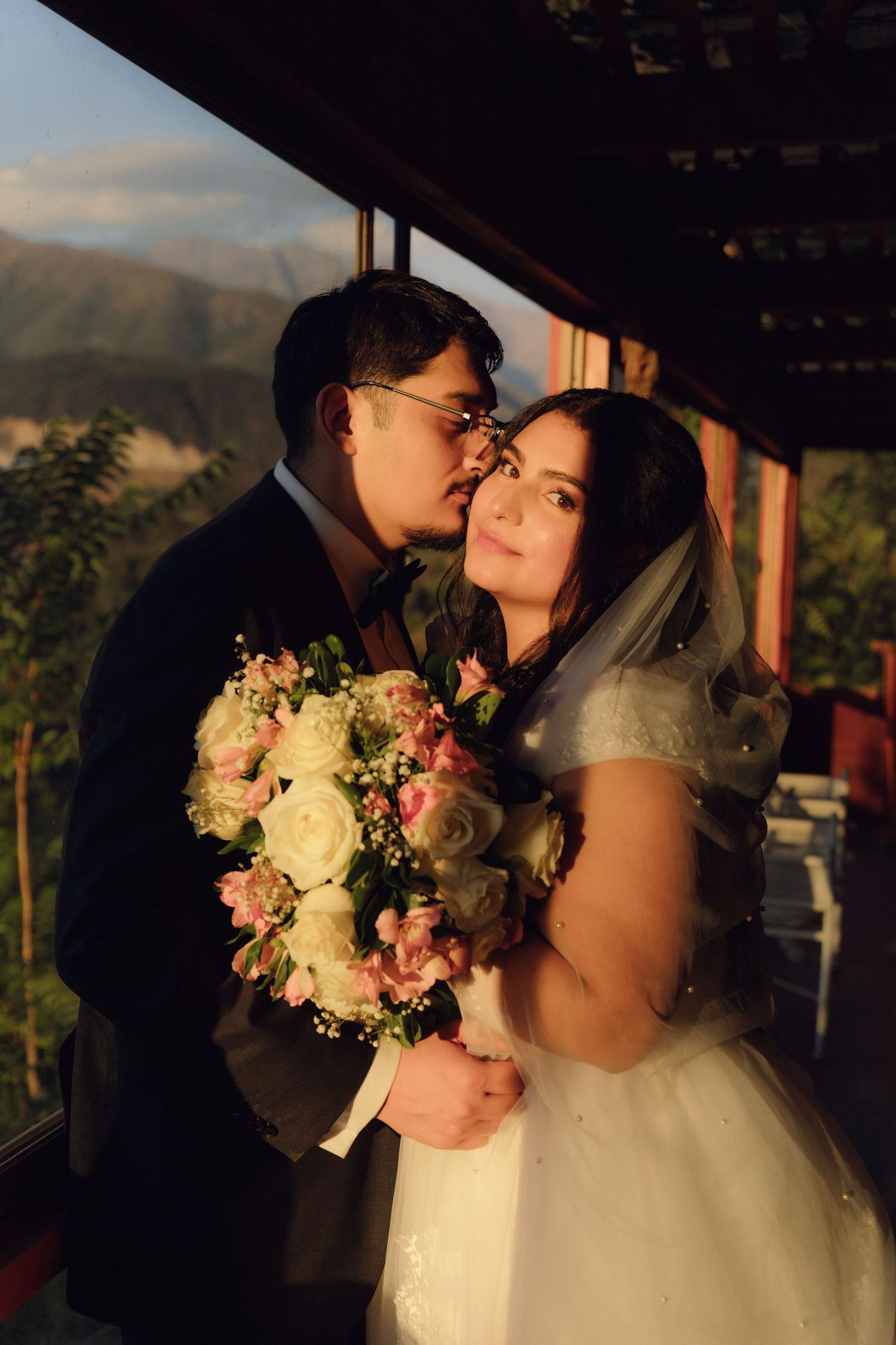 Groom tenderly embraces bride holding white and pink bouquet on balcony with mountain sunset backdrop.