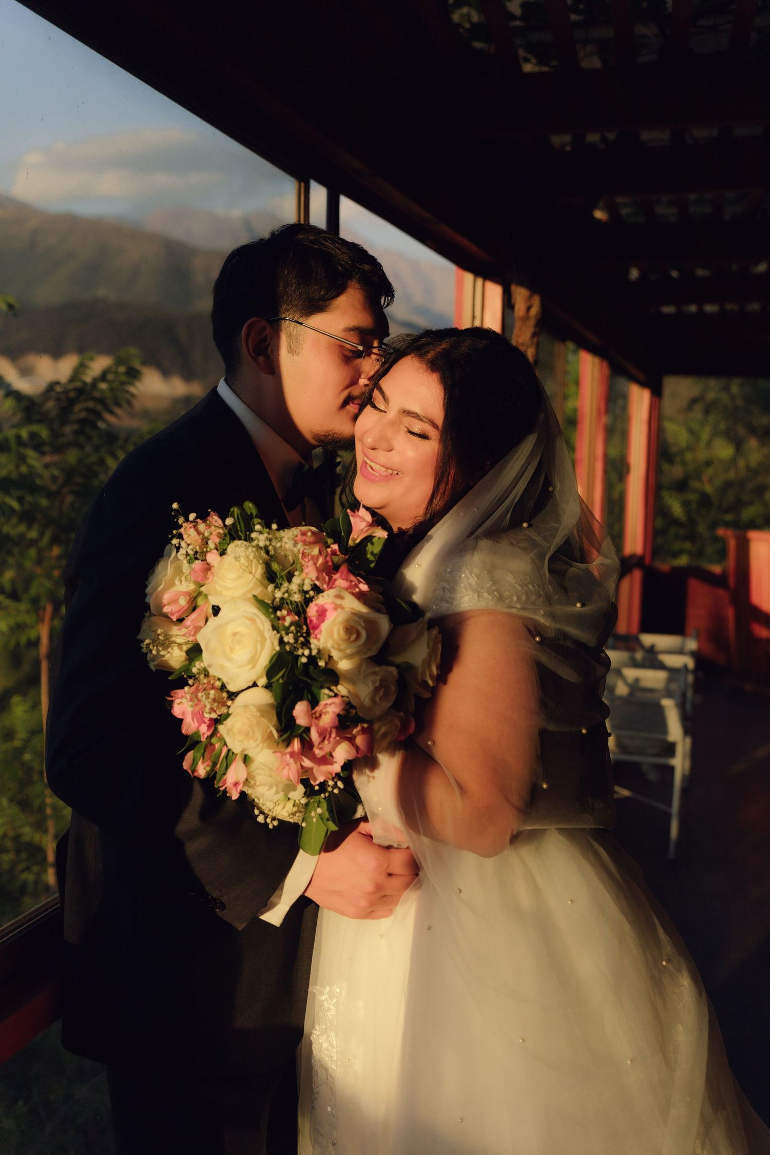 Groom kisses smiling bride on cheek as she holds floral bouquet with mountains at sunset.