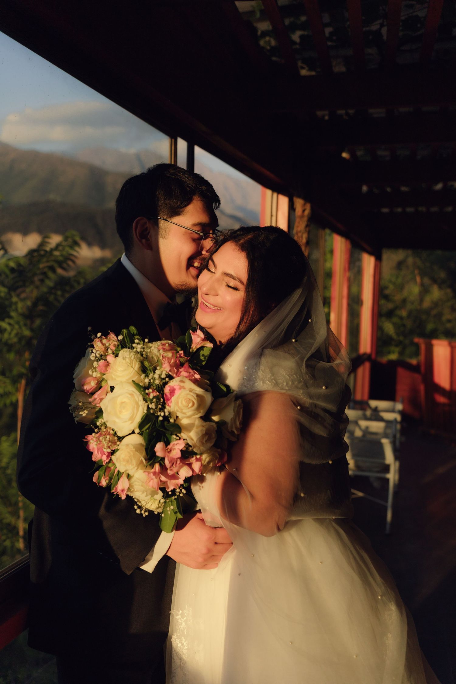 Groom nuzzles bride from behind as she smiles holding bouquet on rustic balcony at sunset.