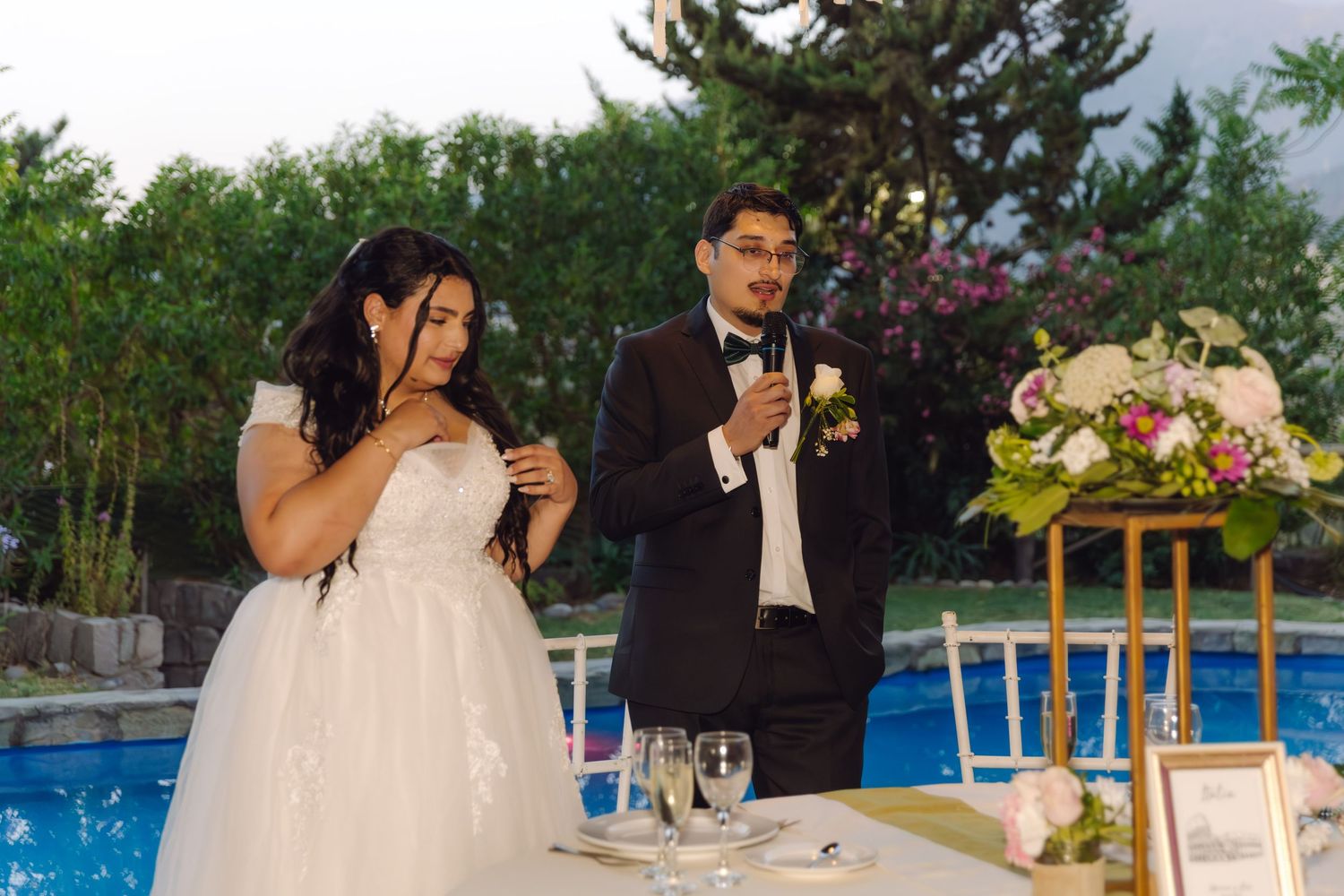 Bride and groom stand together at outdoor wedding reception near pool, groom speaking into microphone.