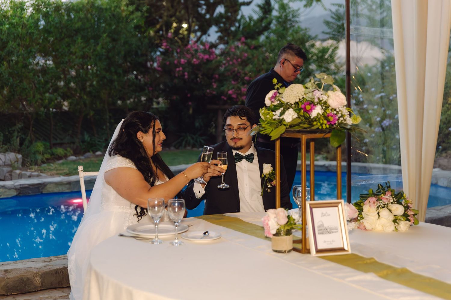 Bride and groom toast champagne glasses at sweetheart table during outdoor poolside wedding reception.