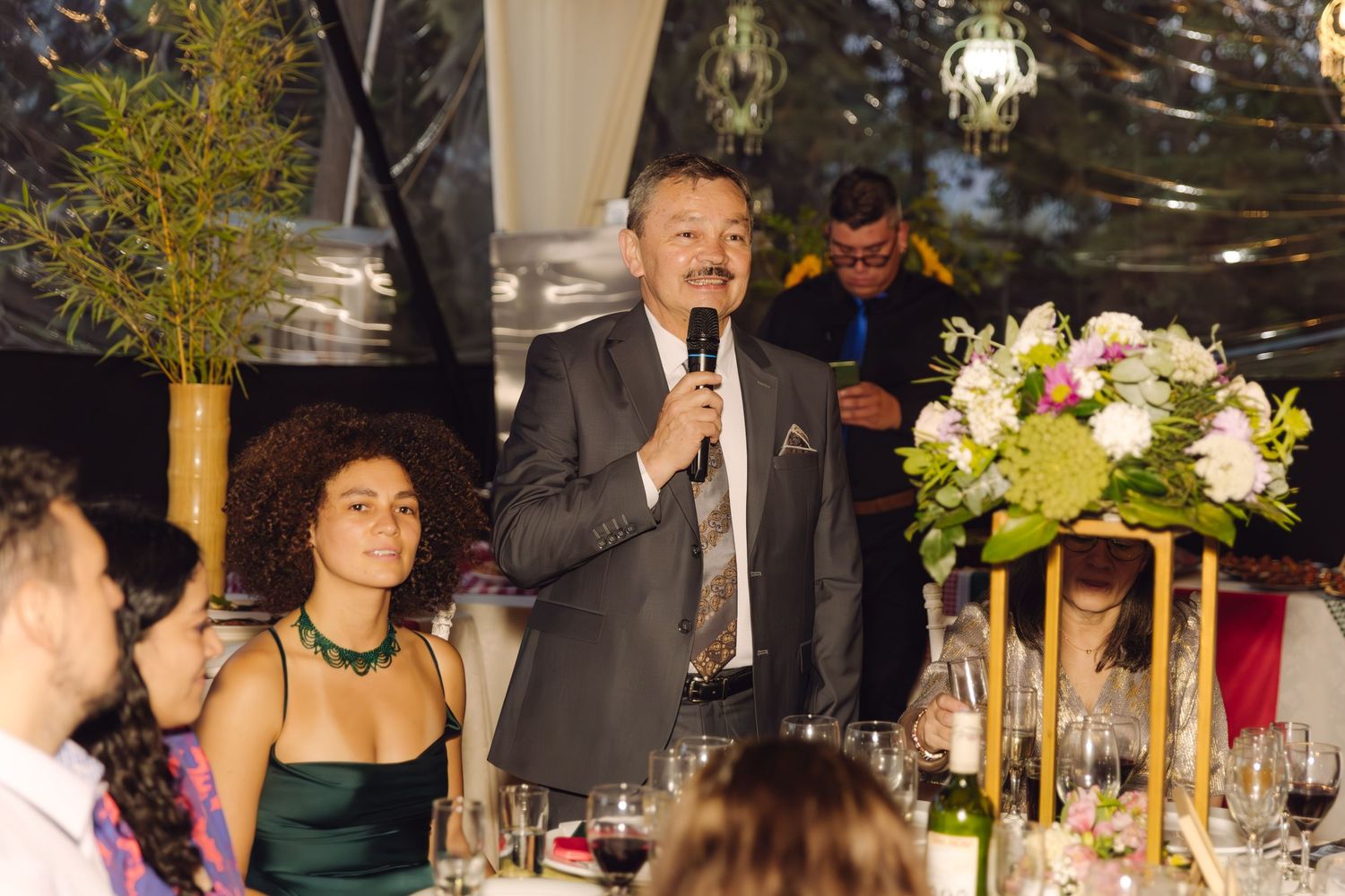 Elegantly dressed man delivers speech into microphone at indoor wedding reception dinner.