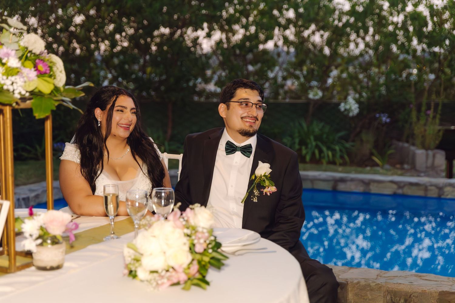Bride and groom sit together at sweetheart table during outdoor poolside wedding reception at dusk.