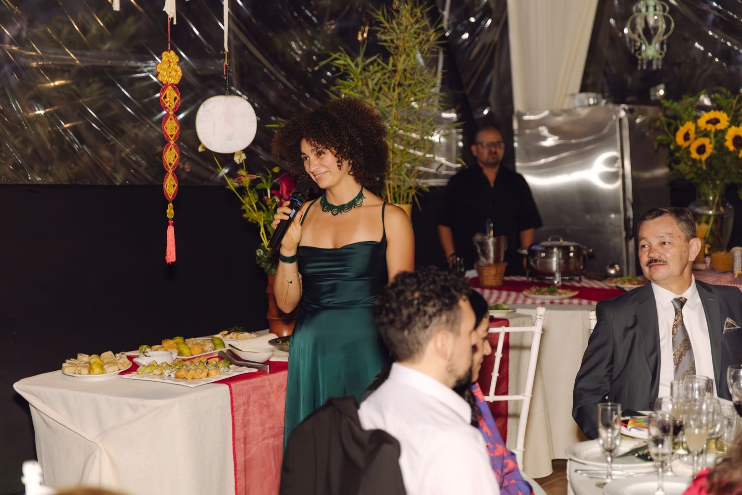 Woman in emerald green dress speaks at wedding reception table with sunflowers and guests nearby.