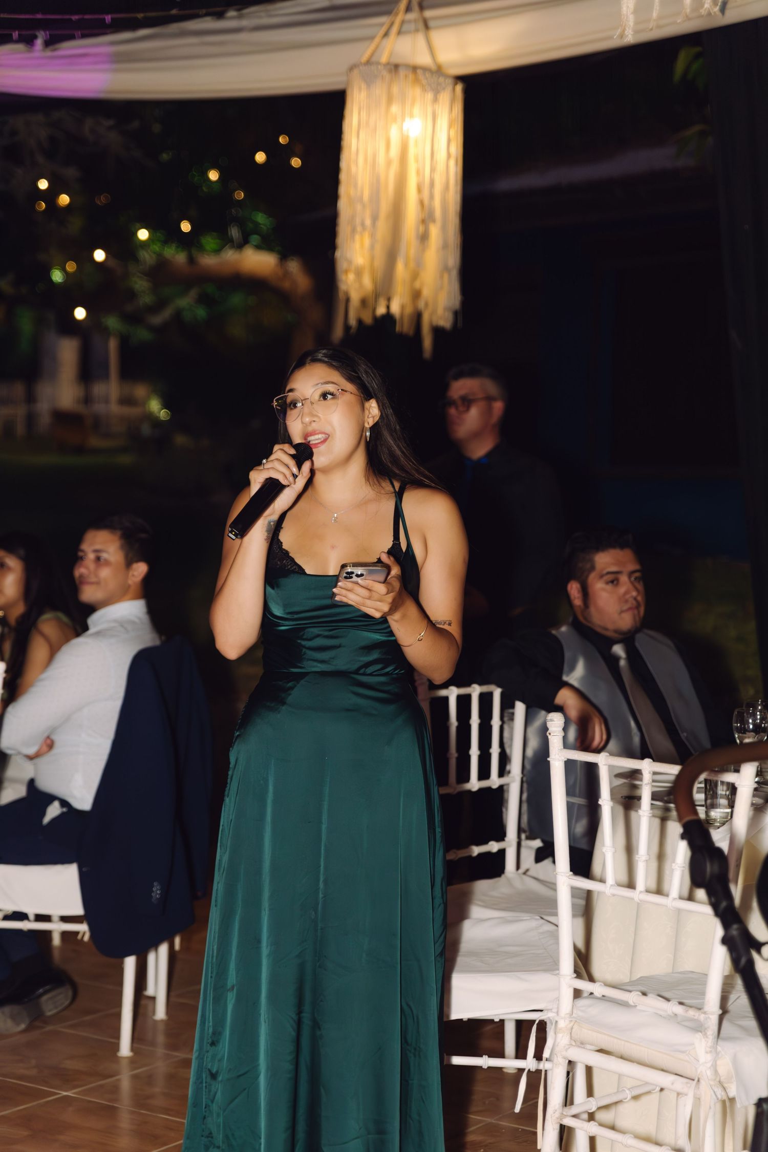 Woman in green satin gown gives speech with microphone at elegant outdoor evening wedding reception.