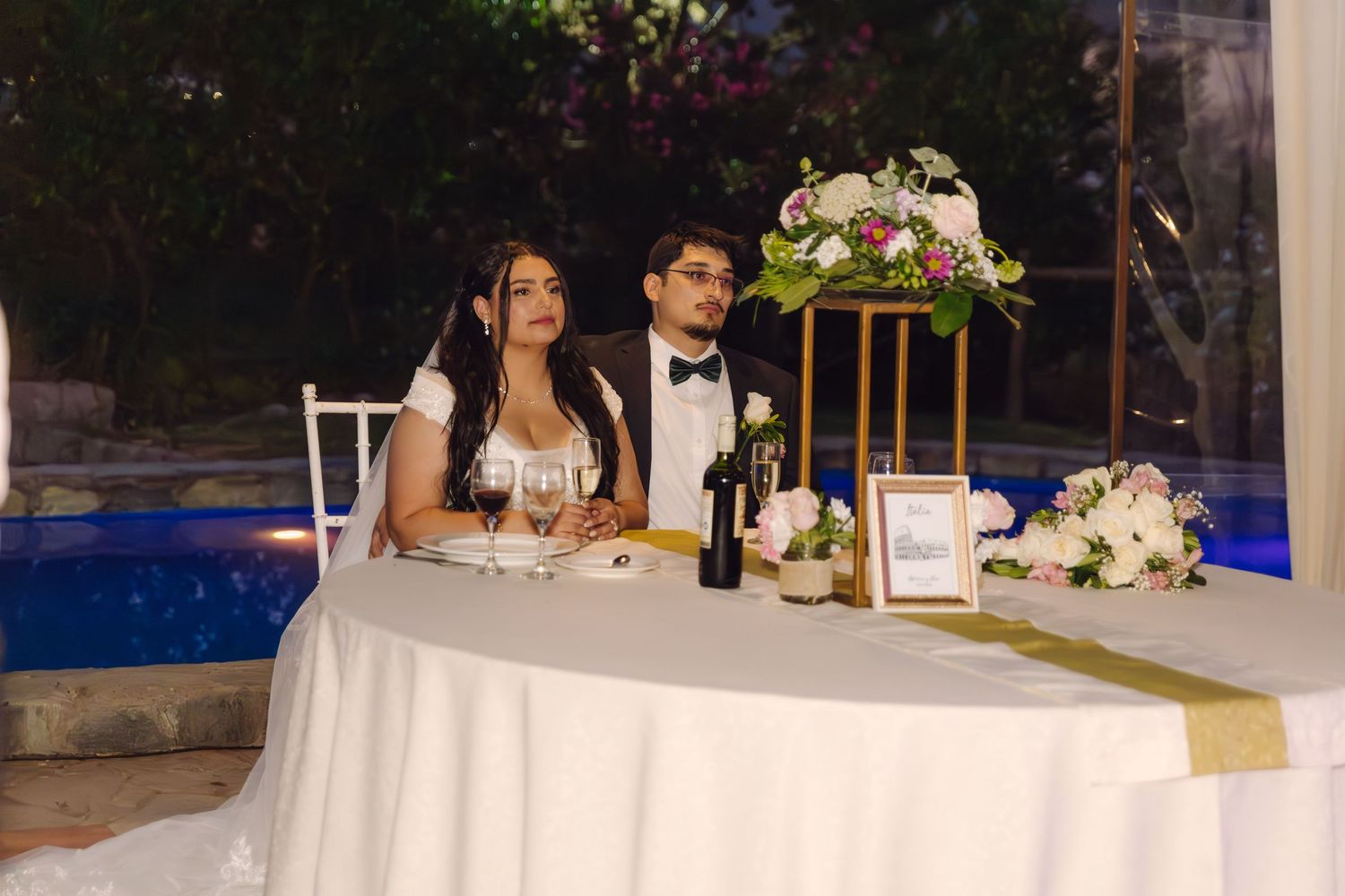 Bride and groom sit at decorated sweetheart table with floral arrangement by illuminated pool at night.