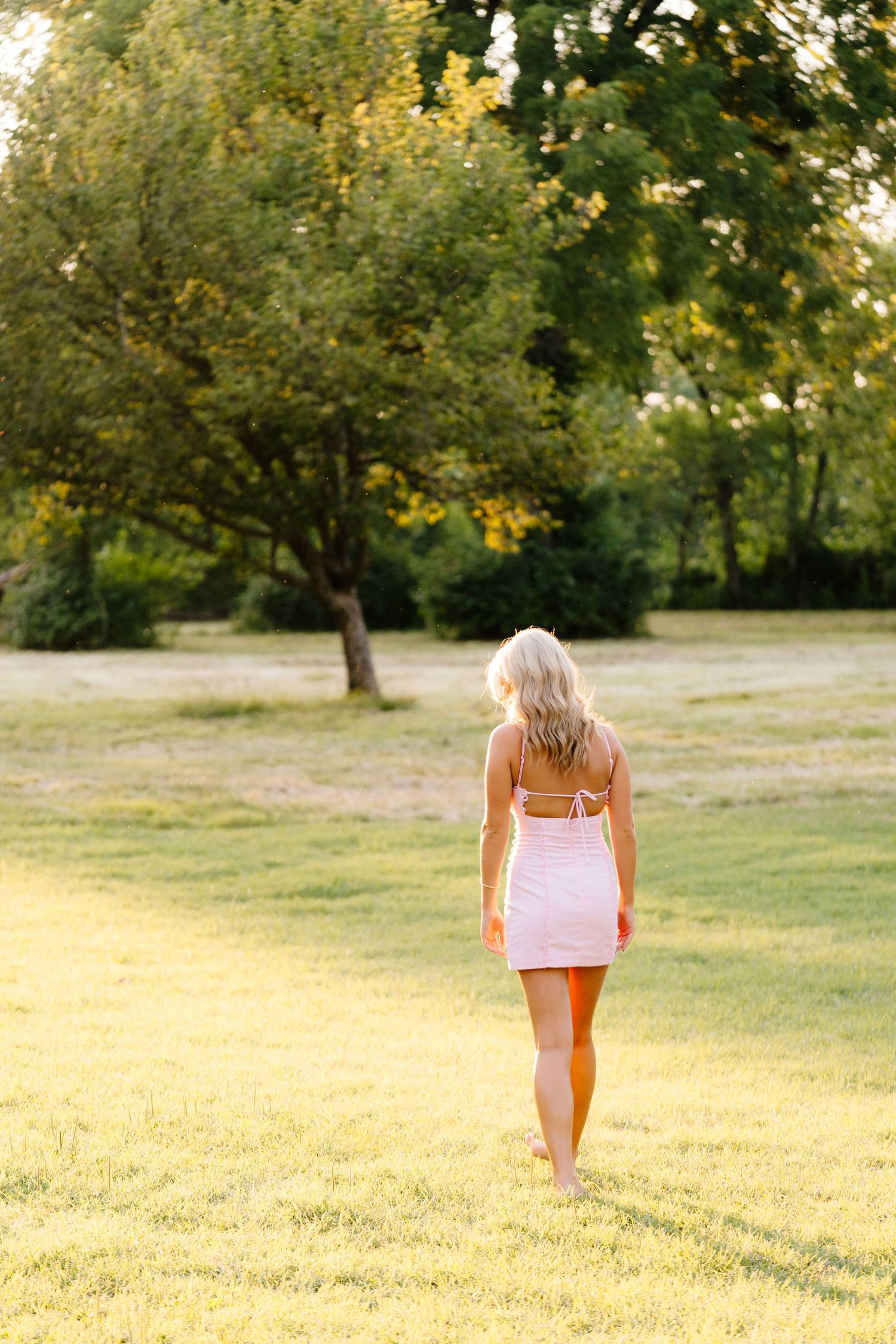 Smiling blonde woman in a pink mini dress standing on green grass with trees in the background on a sunny day.