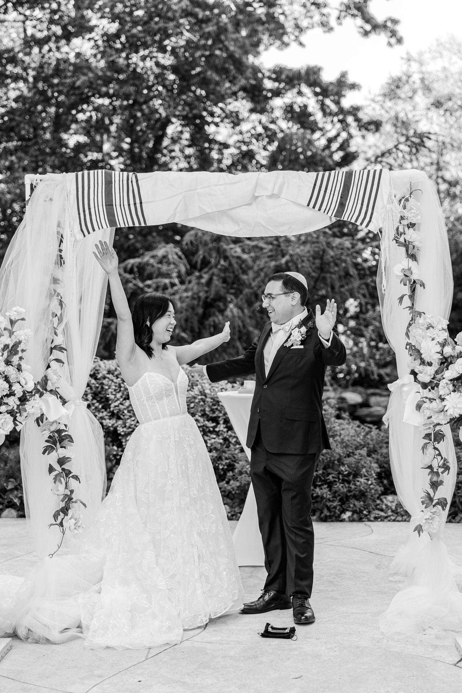 Black and white photo of bride and groom celebrating under floral chuppah at outdoor wedding ceremony.