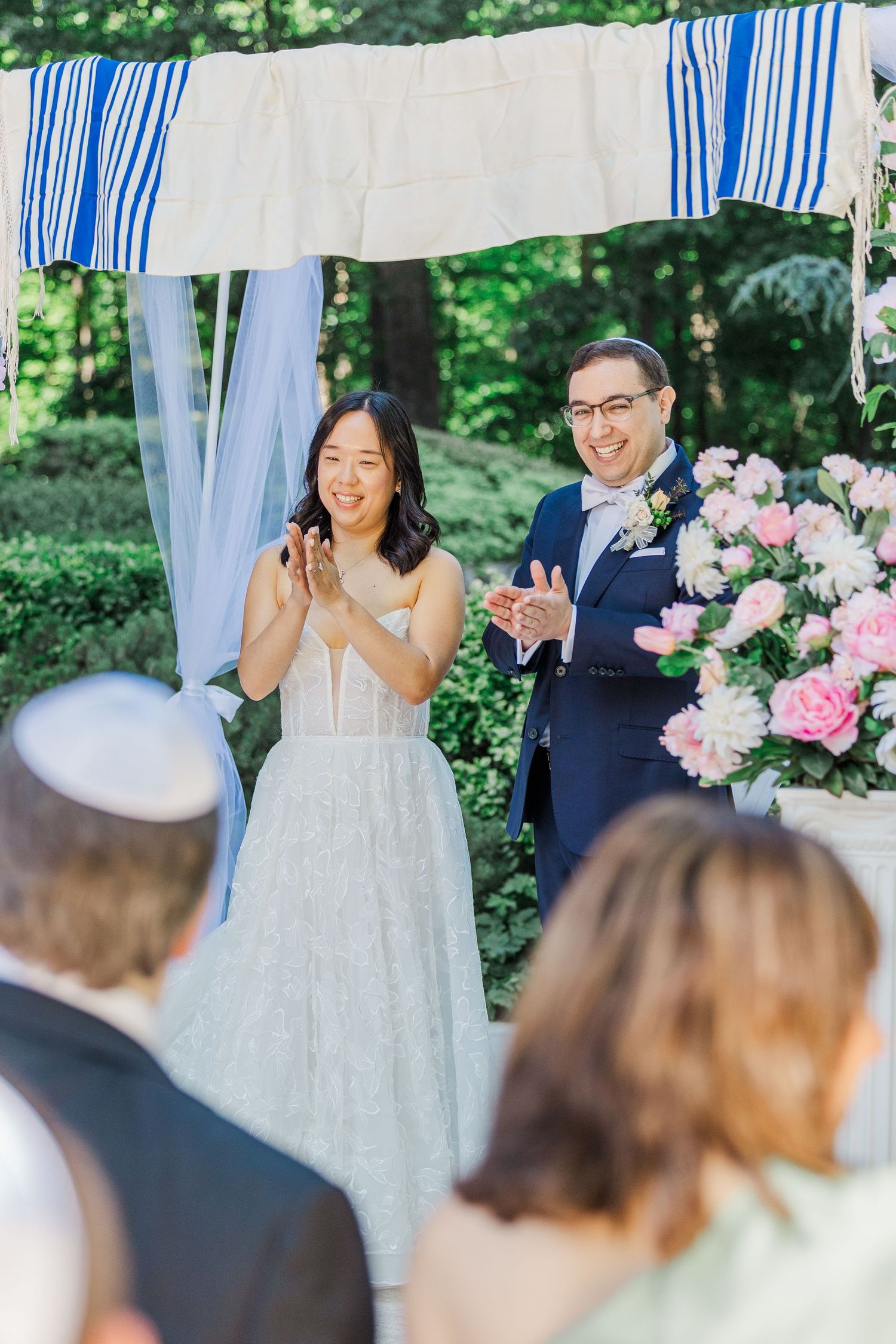 Couple clapping joyfully under a floral chuppah at an outdoor Jewish wedding ceremony surrounded by lush greenery.