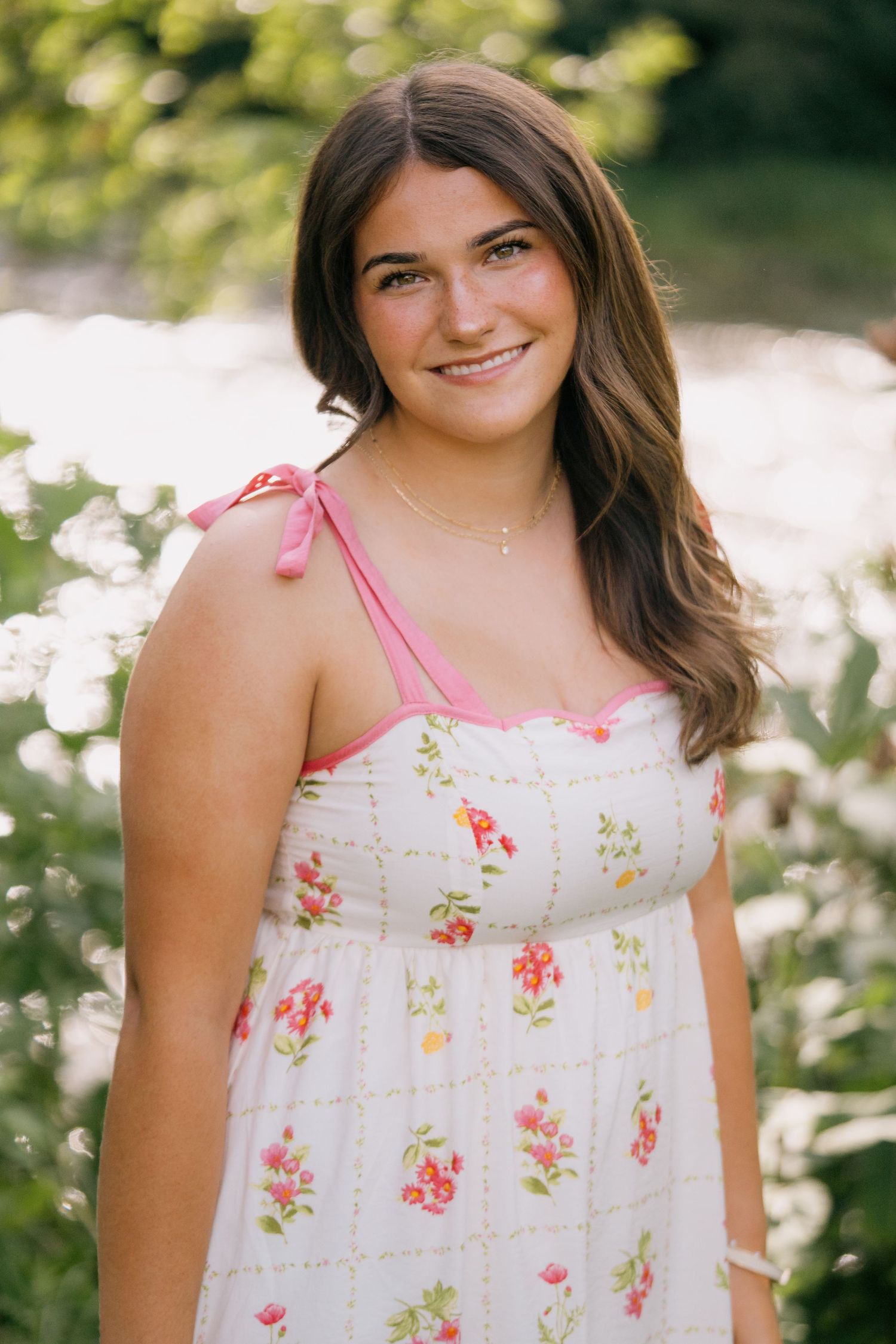 Young woman in floral sundress smiling outdoors near water with lush green foliage in soft natural light.