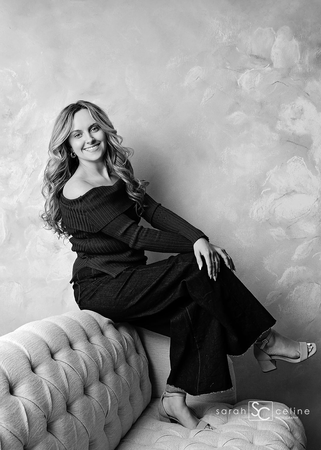 Black and white studio portrait of a smiling woman in dark outfit seated on tufted sofa, by Sarah Celine Photography.