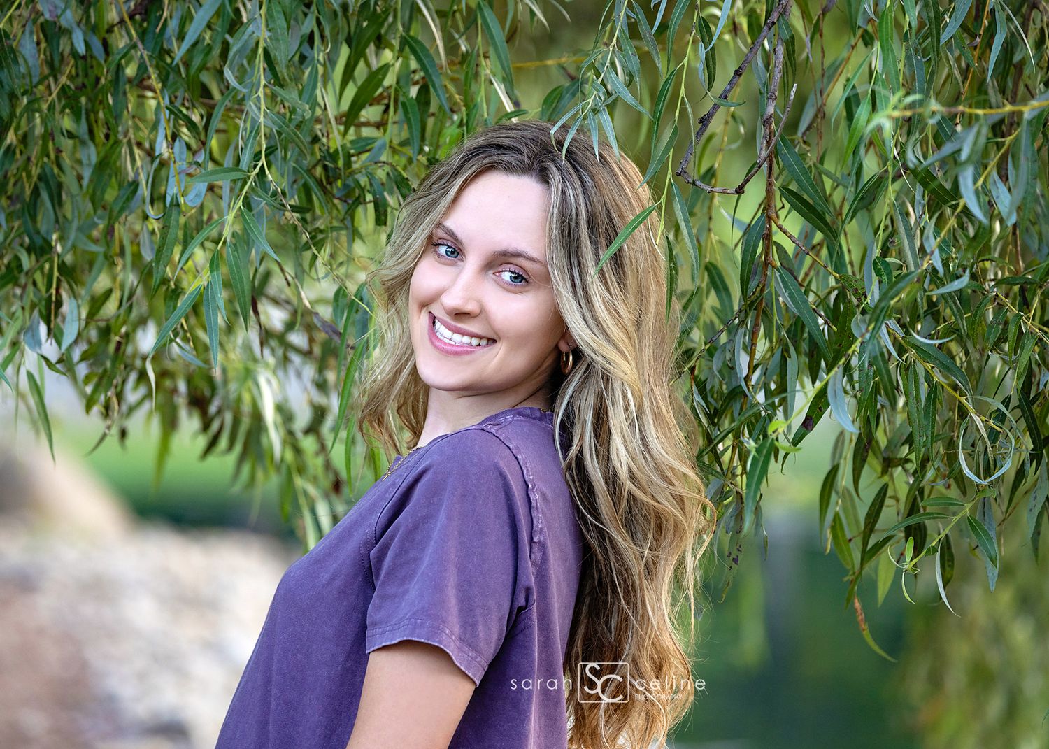 Young woman with long wavy blonde hair smiling under weeping willow branches outdoors.