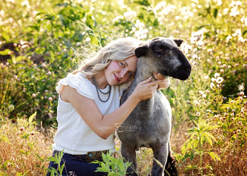Smiling blonde woman hugging a large dark-colored dog in a sunny wildflower field.
