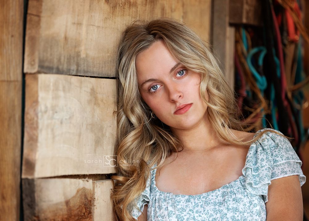 Young blonde woman with blue eyes in a floral top leaning against rustic wooden wall.