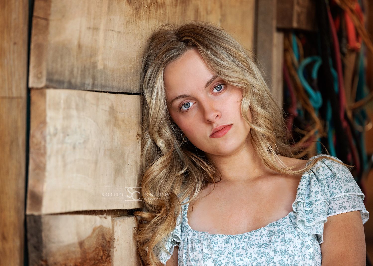 Young blonde woman with blue eyes in a floral top leaning against rustic wooden wall.