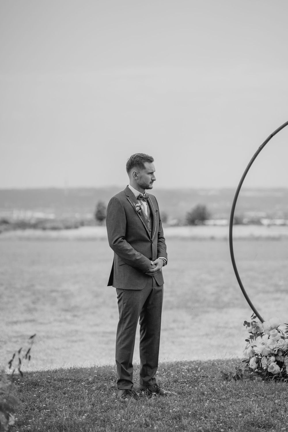 Groom in a gray suit standing by a circular floral arch at an outdoor wedding ceremony.