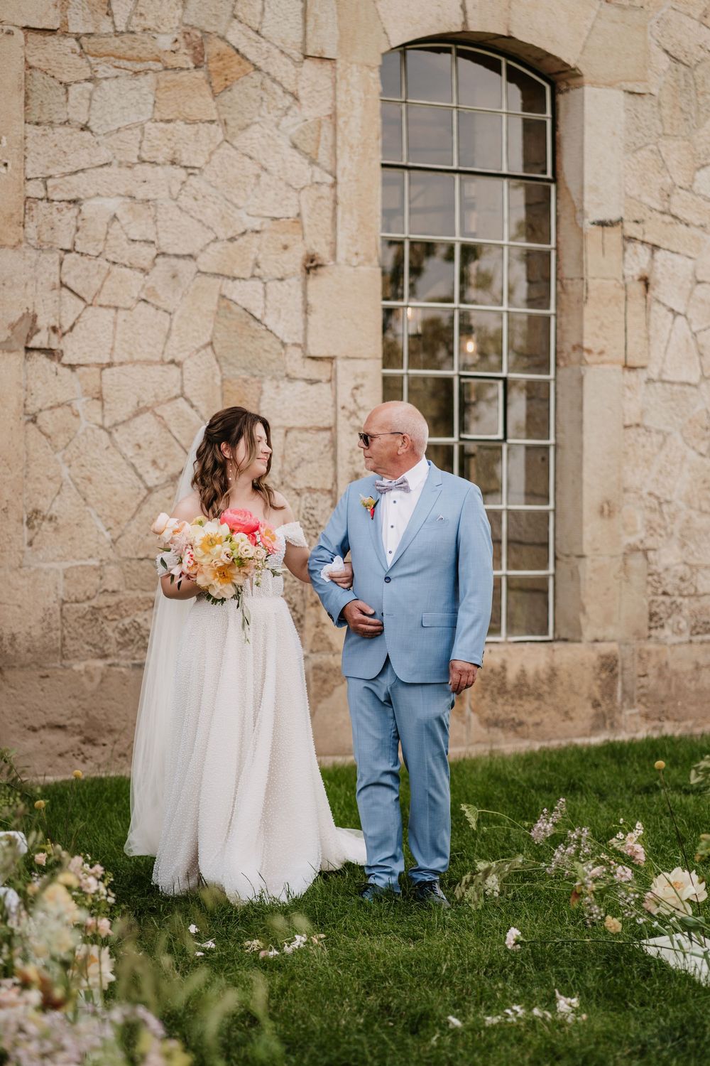 Bride in white gown holding bouquet walks with man in light blue suit outside stone building.