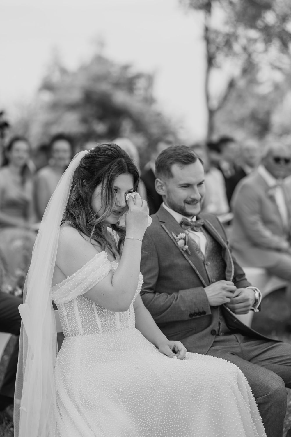 Bride wiping tears while groom smiles during an outdoor wedding ceremony.