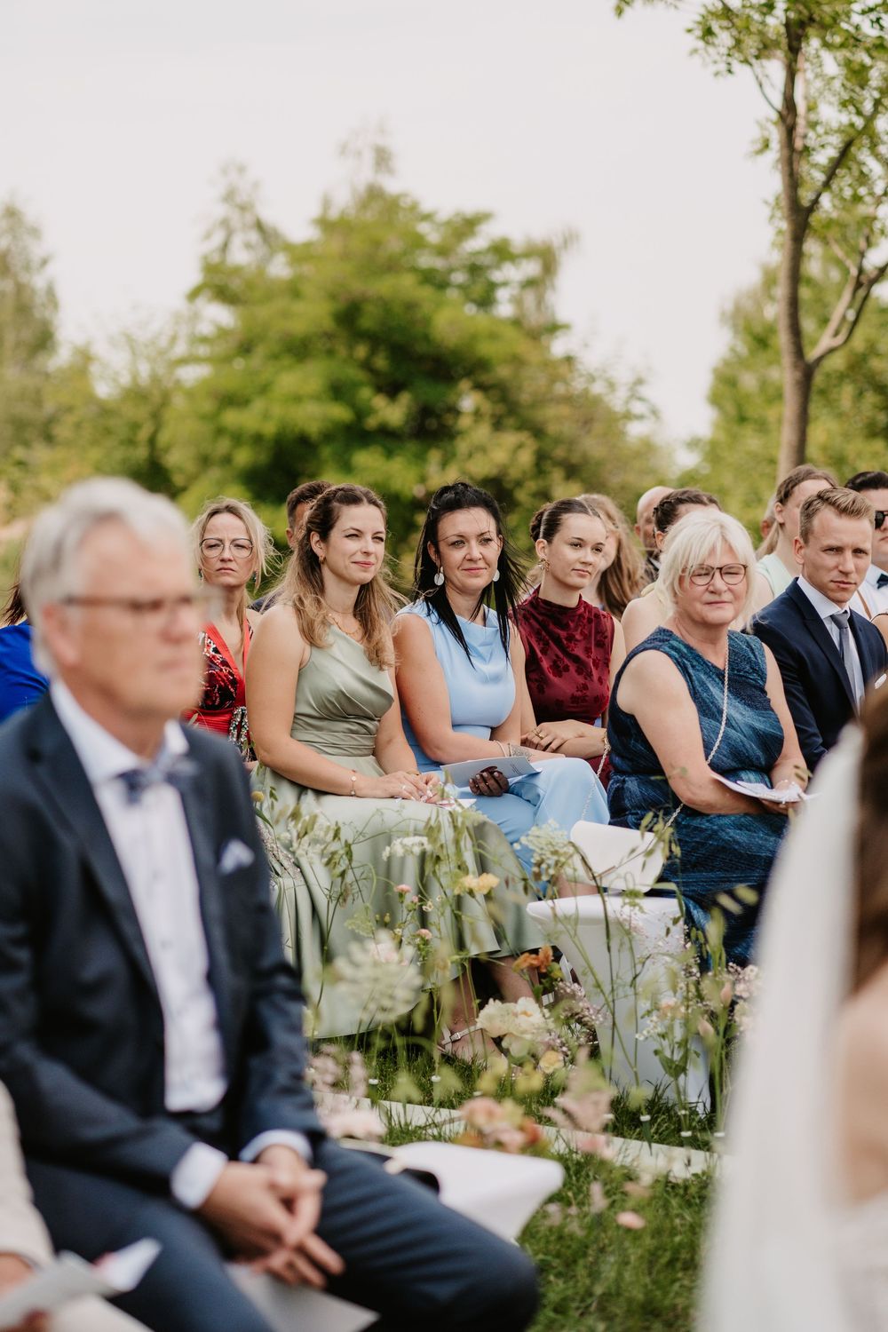 Wedding guests seated outdoors watching a ceremony, surrounded by lush green trees and floral arrangements.