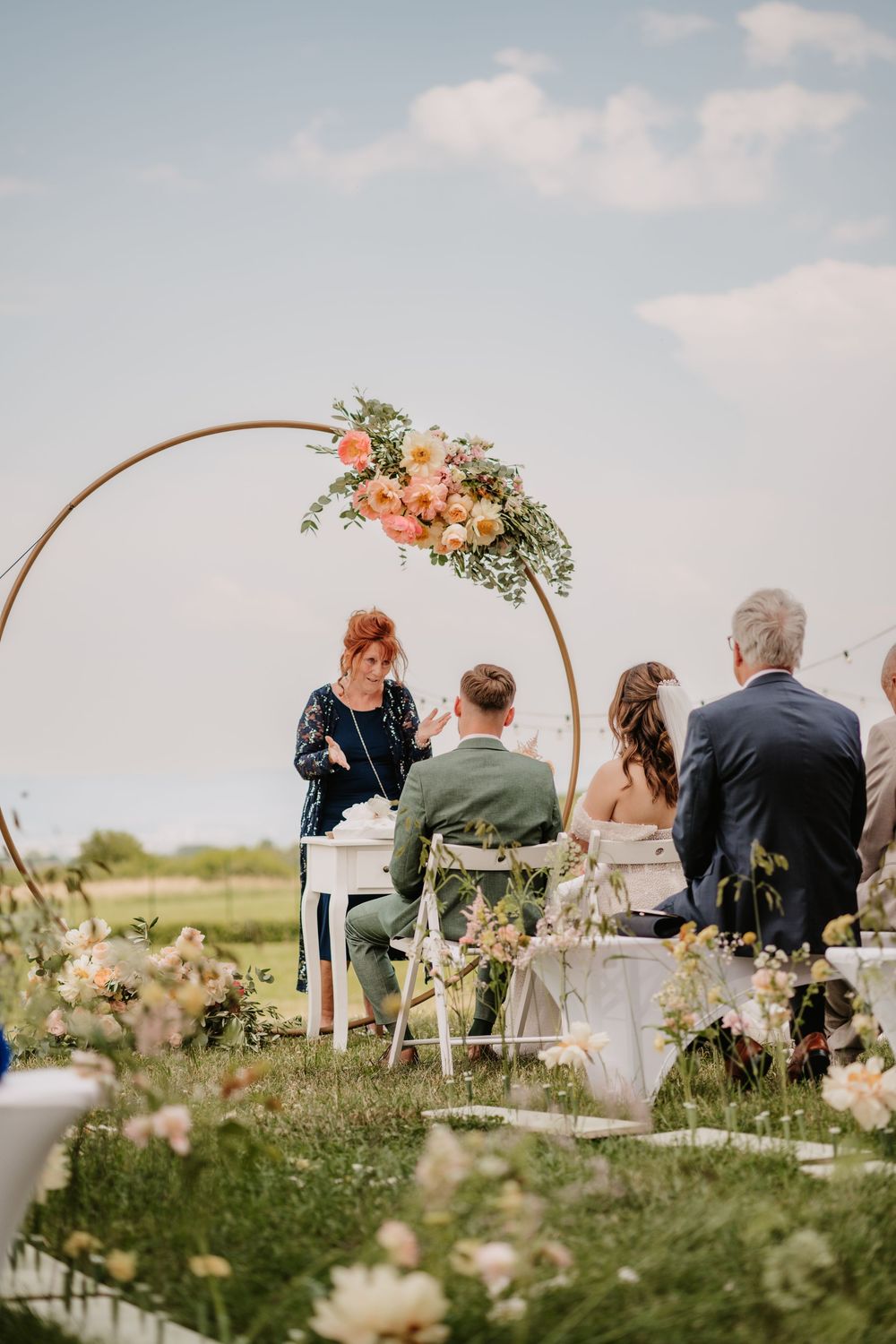Officiant speaking at an outdoor wedding ceremony with a circular floral arch backdrop.