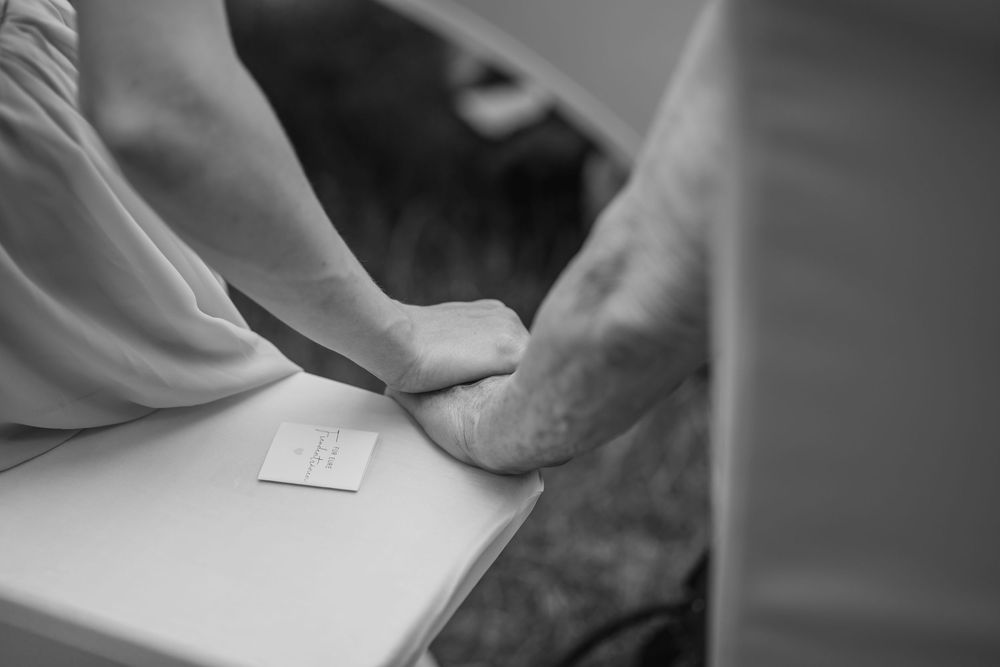 Black and white photo of two people holding hands on a white surface with a small card nearby.
