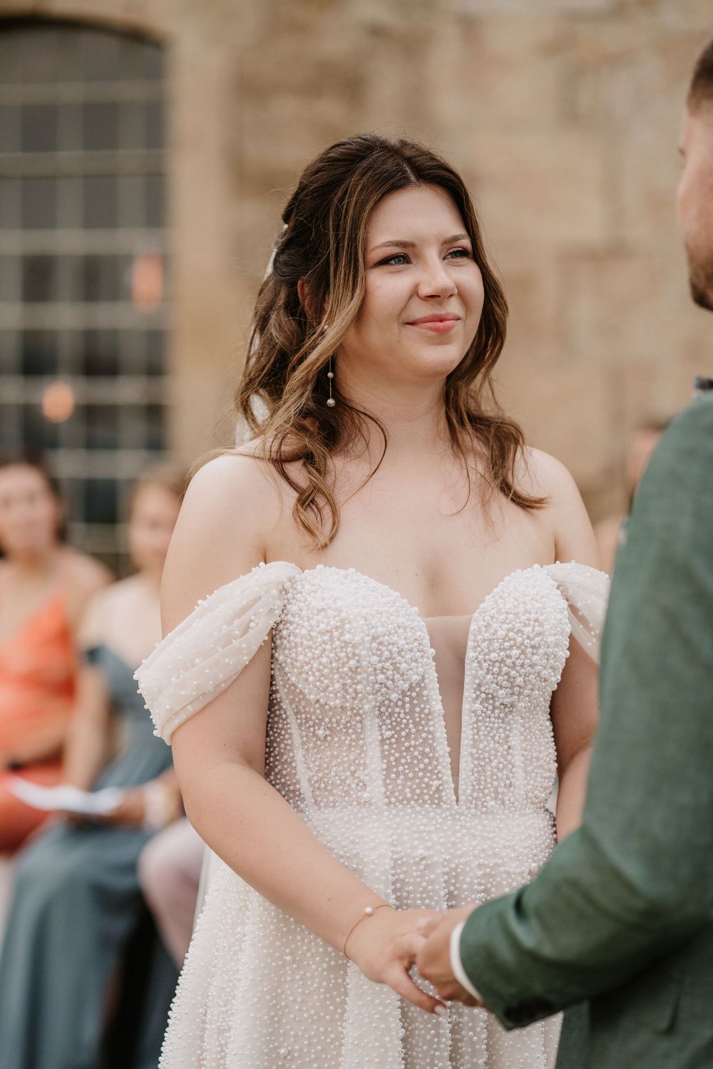 Bride in pearl off-shoulder wedding gown smiling while holding hands with groom during outdoor ceremony.