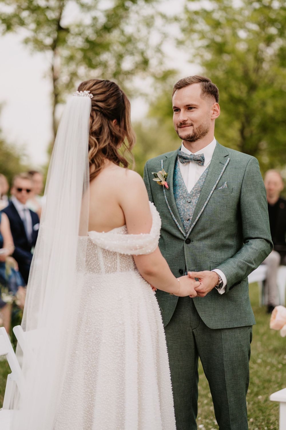 Groom in green suit holds bride's hands during outdoor wedding ceremony with guests seated behind.