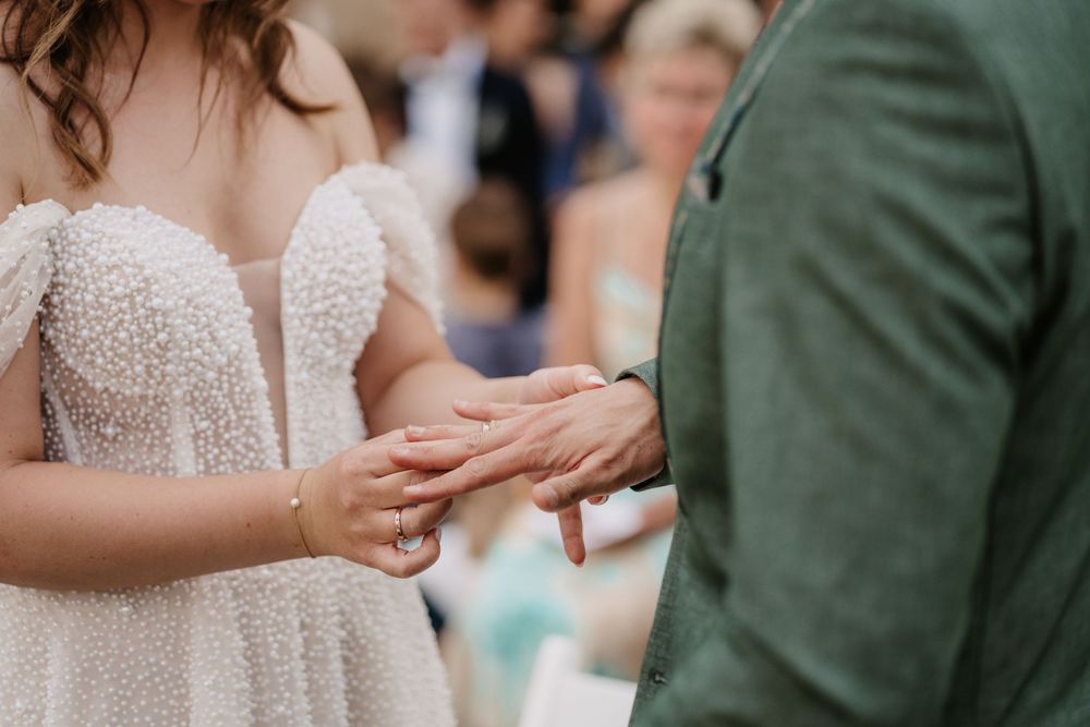 Bride placing a wedding ring on groom's finger during outdoor ceremony, wearing beaded white gown.