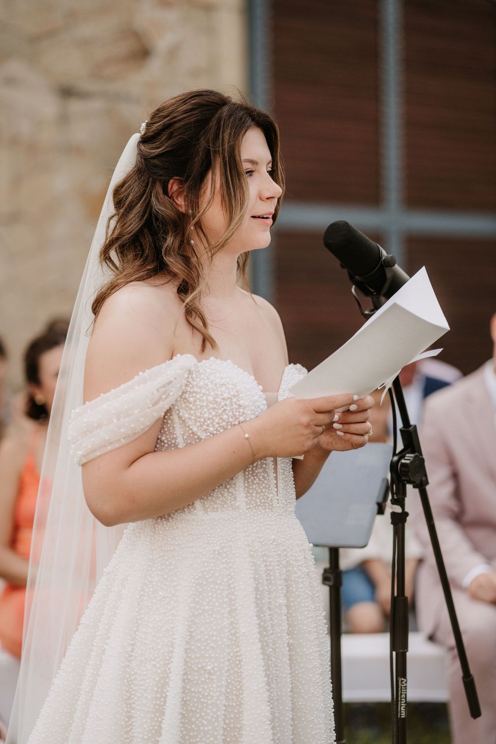 Bride in a pearl off-shoulder gown reading vows at a microphone during outdoor ceremony.