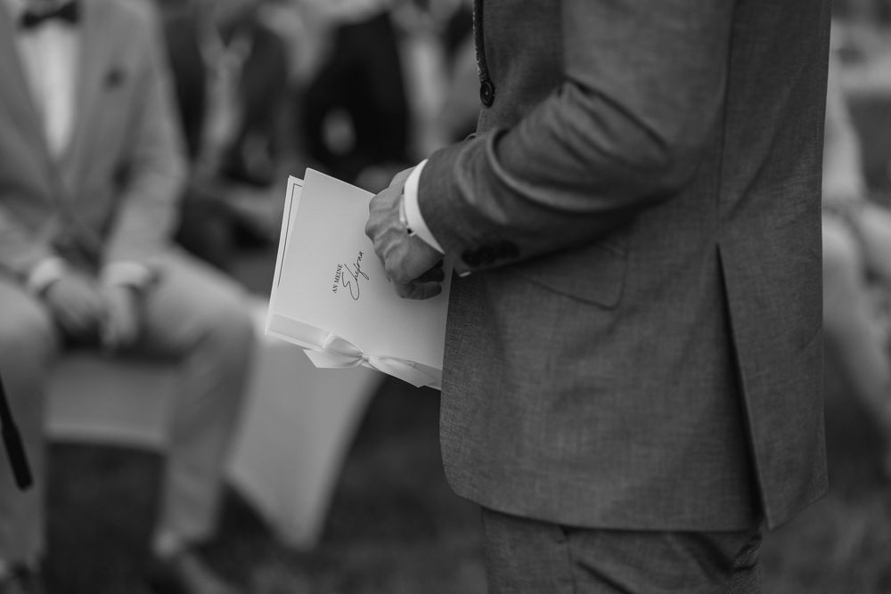 Black and white photo of a groom in a gray suit holding wedding ceremony papers outdoors.